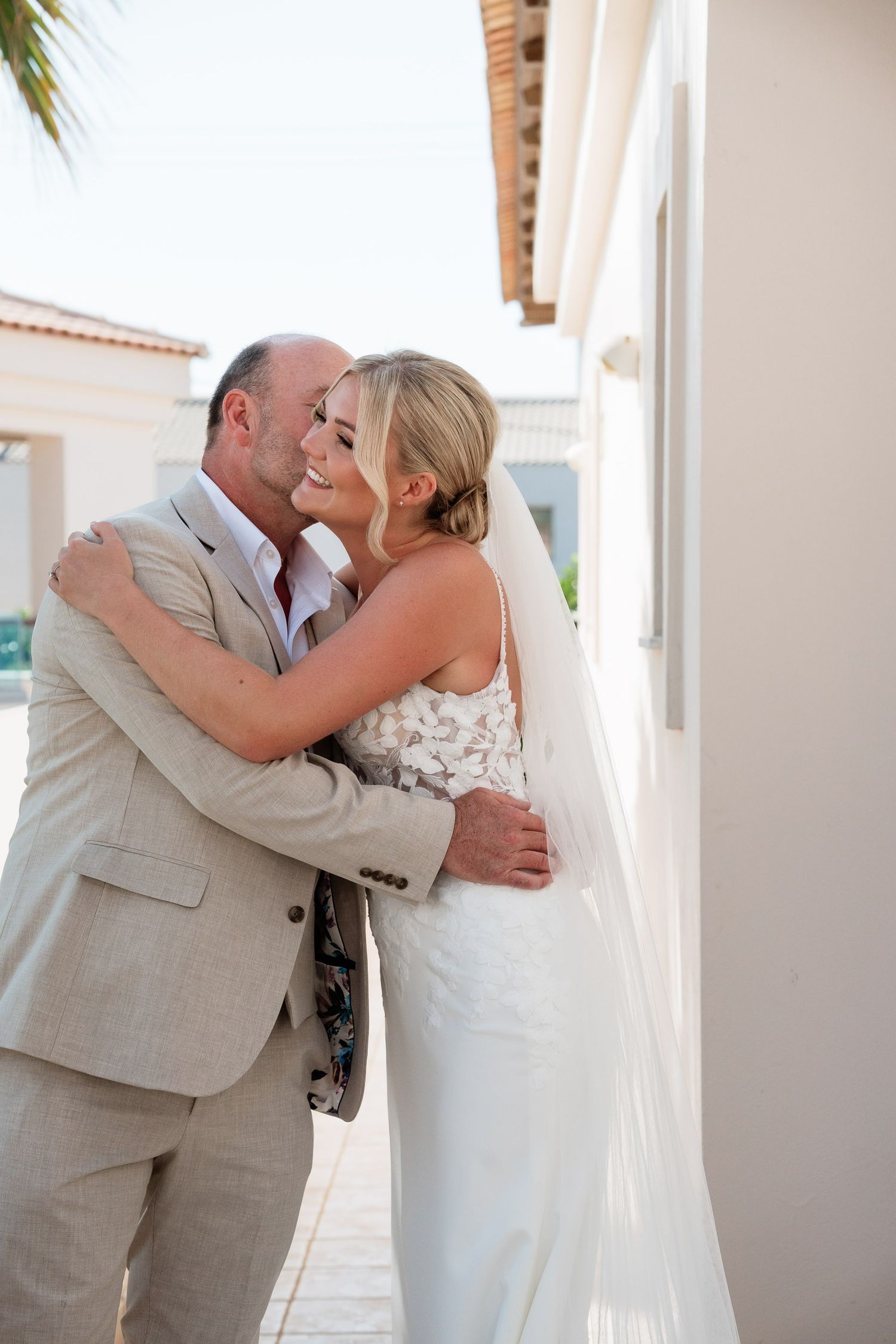 A couple shares a tender moment on their wedding day in front of a white stucco building with Spanish tile roof.