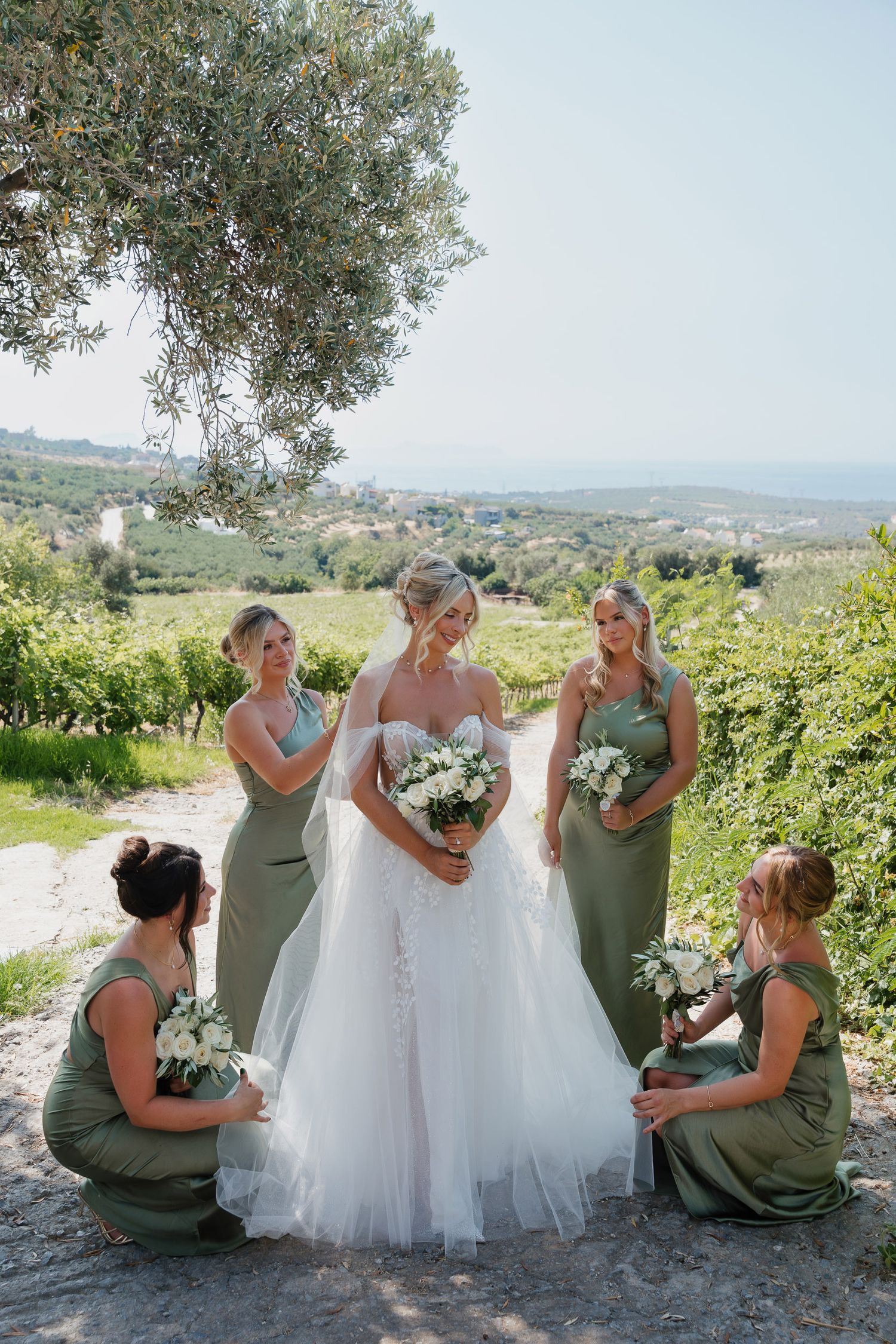 A wedding party in sage green dresses and white gown pose together in a scenic Italian countryside vineyard setting.