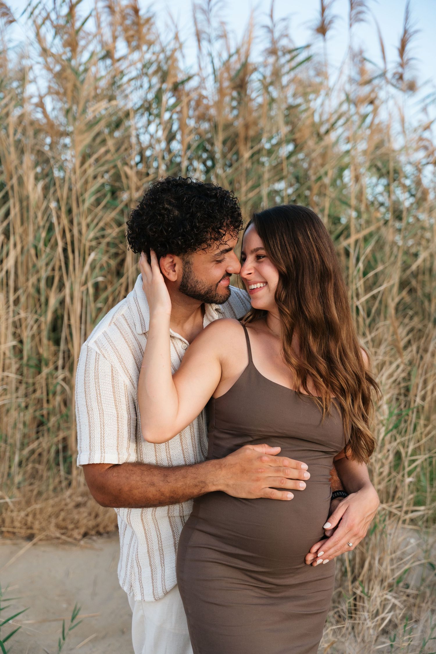 A romantic maternity photoshoot shows an intimate couple embracing among tall reeds on a beach at sunset.
