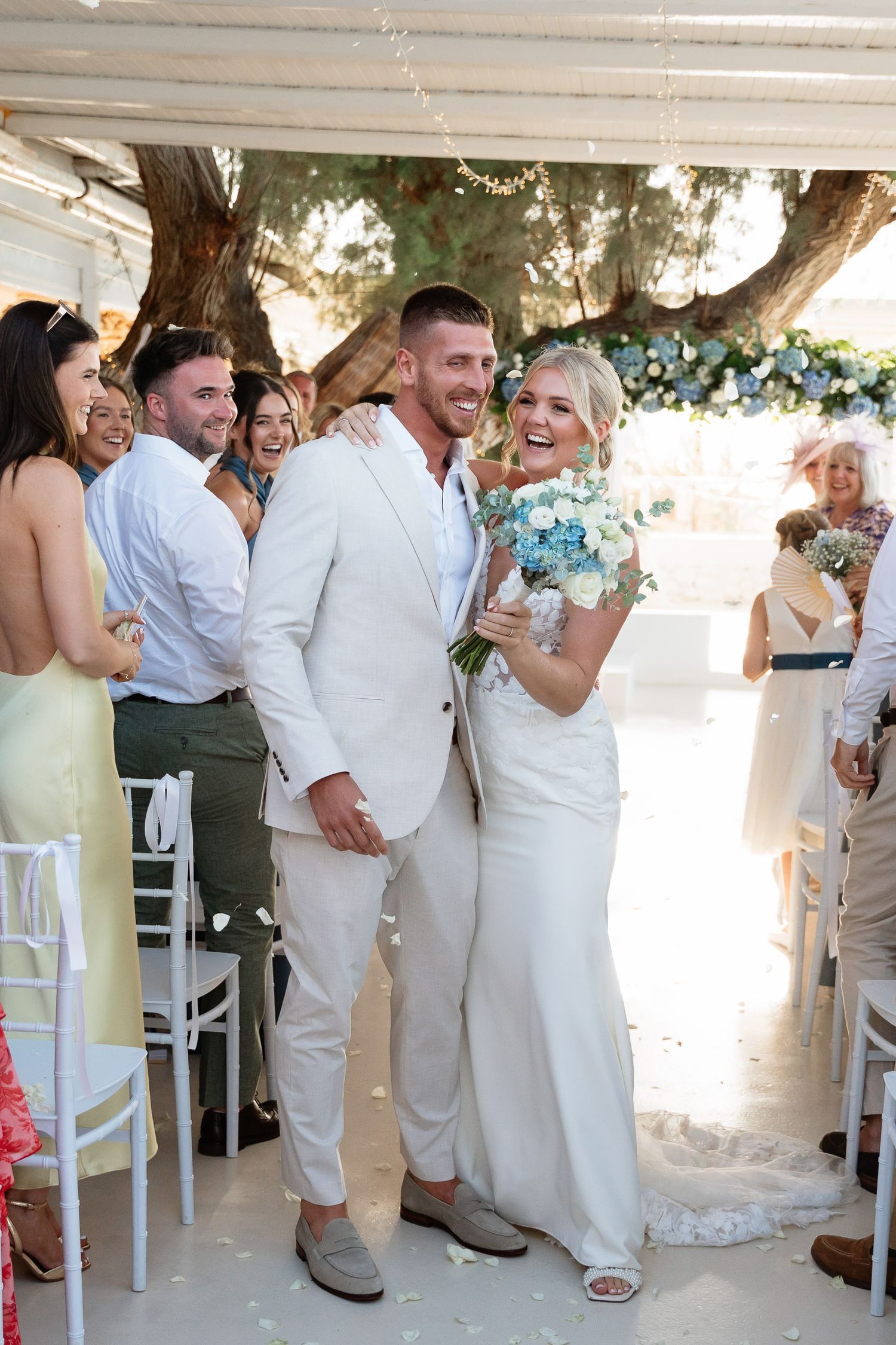 Wedding guests in white and pastel attire celebrate and laugh together during an outdoor reception ceremony.
