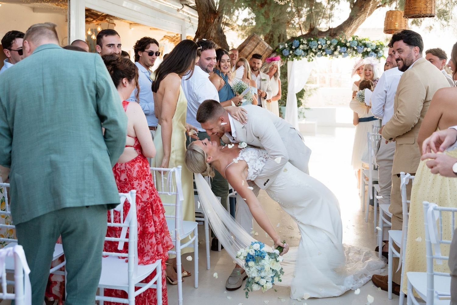 Wedding guests laugh and celebrate during a candid moment at an outdoor reception with white chairs and floral decor.