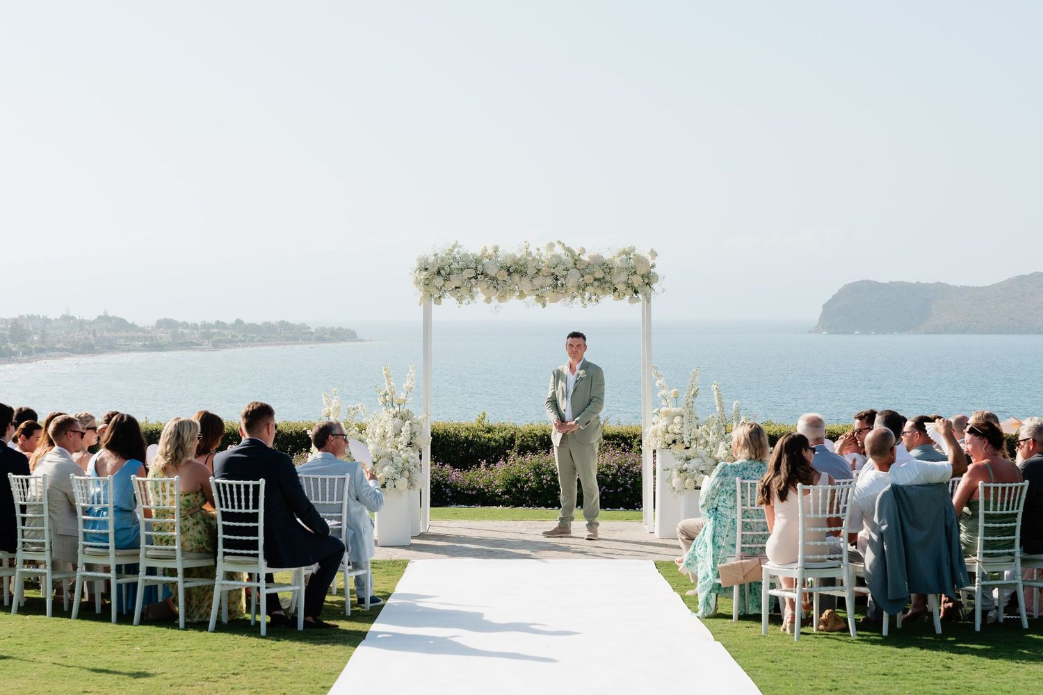 A scenic outdoor wedding ceremony setup with white chairs and floral arch overlooking the ocean on a bright sunny day.
