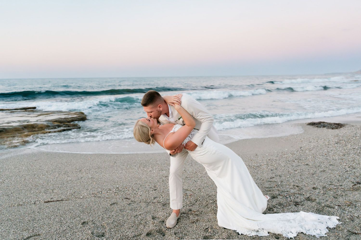 A couple in wedding attire poses romantically on a scenic beach at sunset with waves crashing behind them.