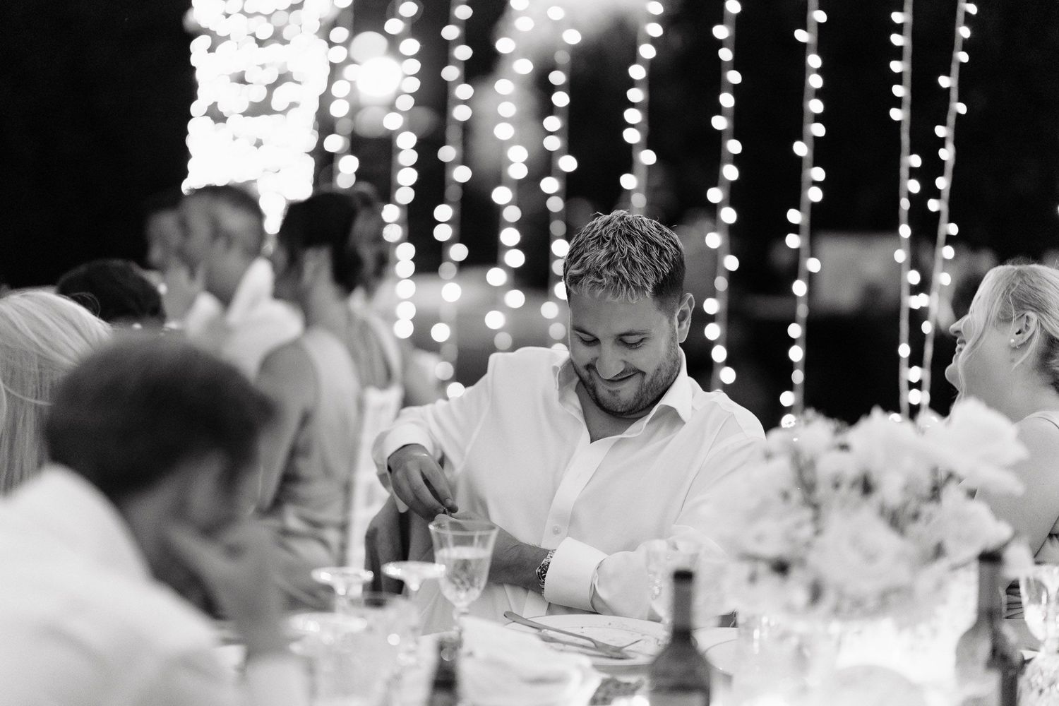 Wedding guests laugh and celebrate at a reception under twinkling string lights during an evening celebration.