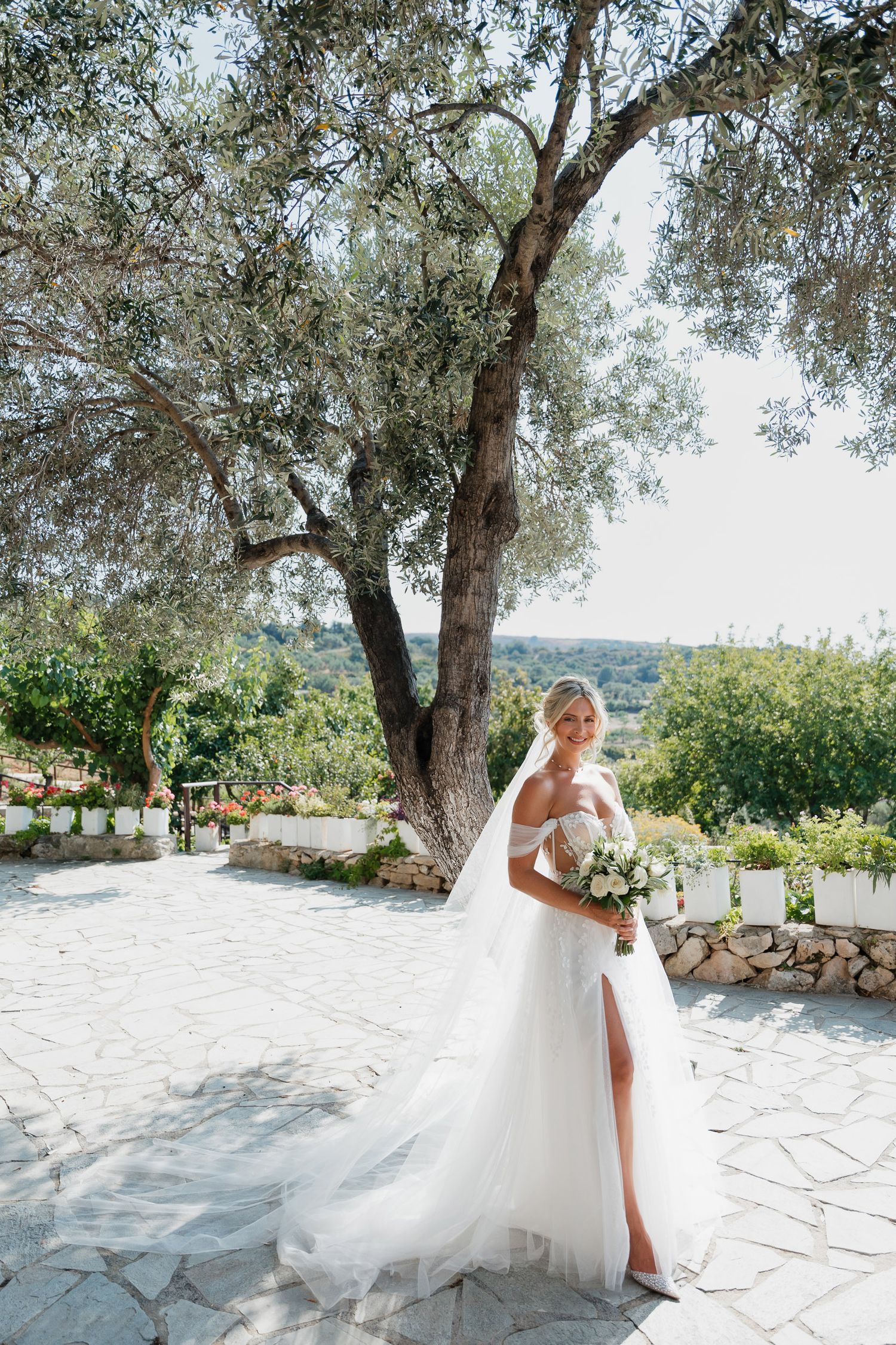 A bride in a flowing white gown twirls gracefully on a stone patio surrounded by olive trees in a Mediterranean setting.