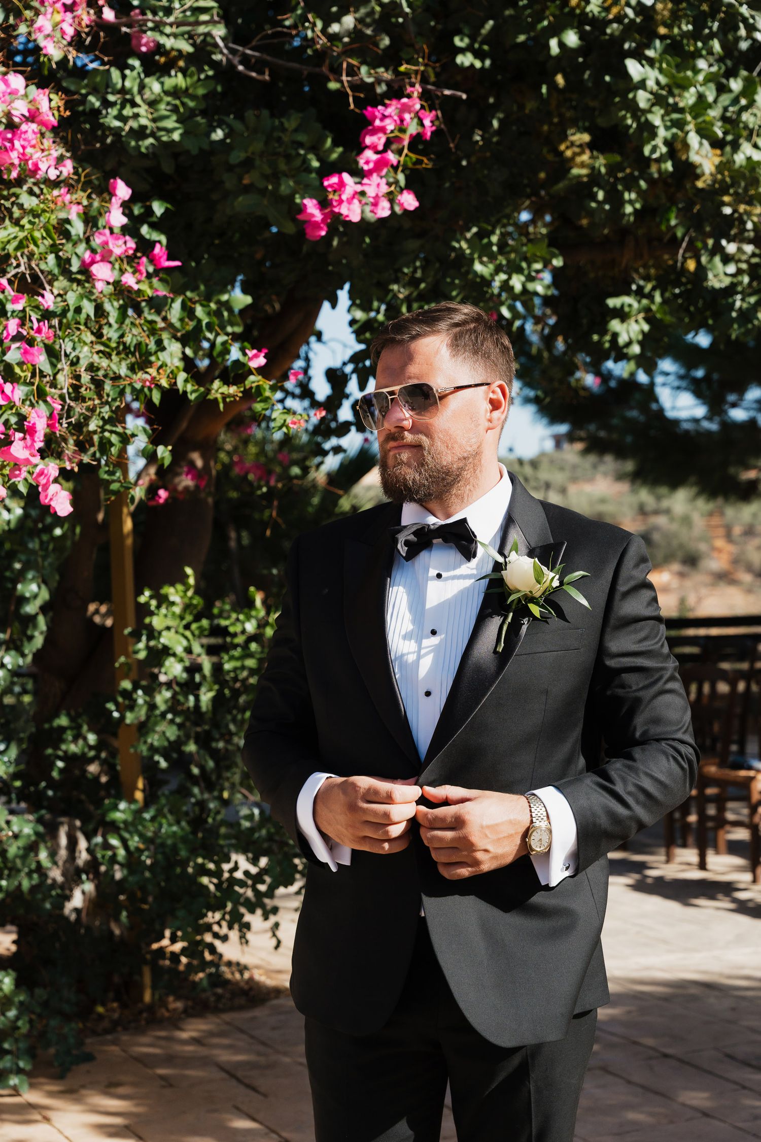Groom in black tuxedo and sunglasses reading vows under pink bougainvillea flowers at outdoor wedding ceremony.
