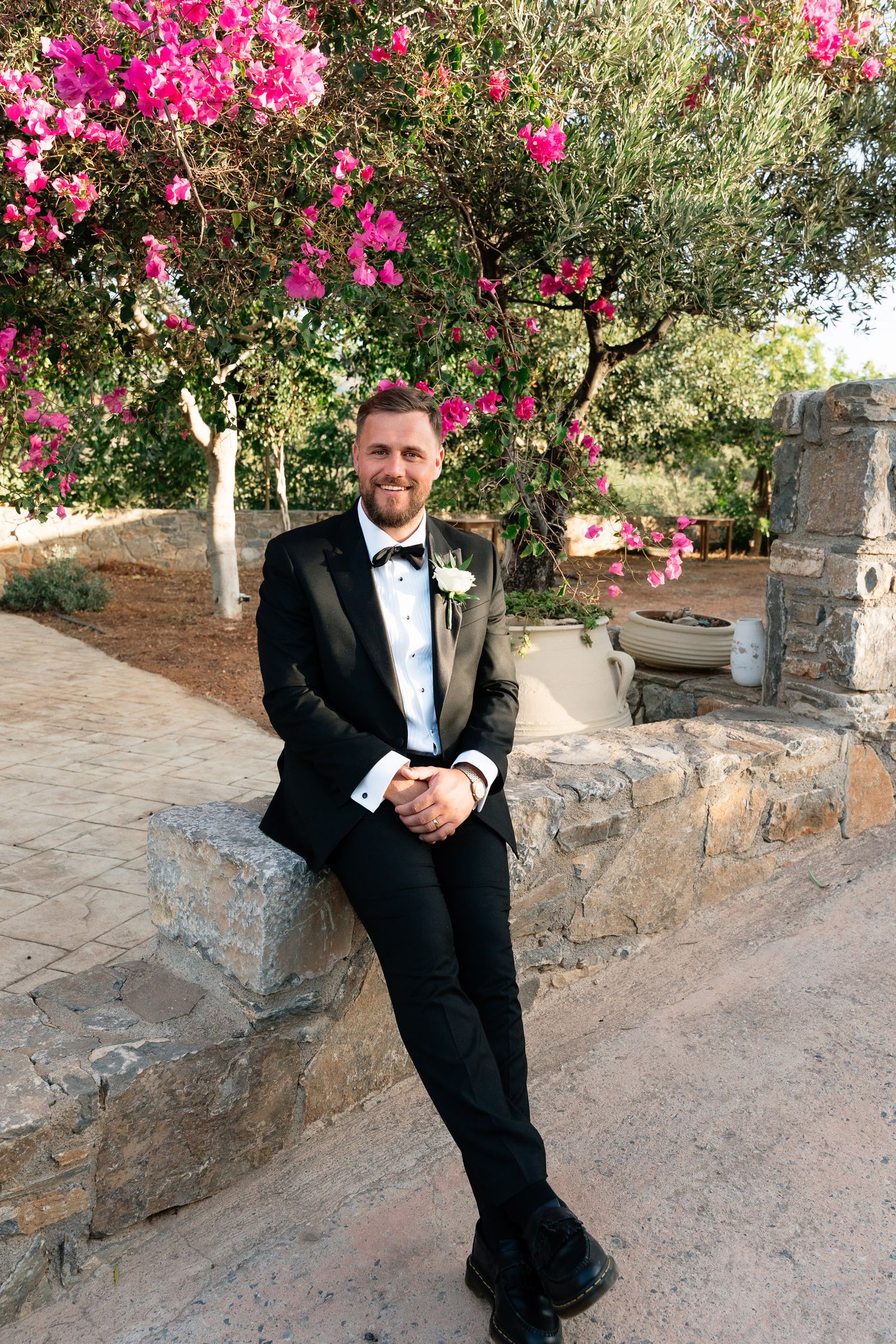 A person in a black tuxedo poses against a stone wall with vibrant pink bougainvillea flowers in a Mediterranean setting.