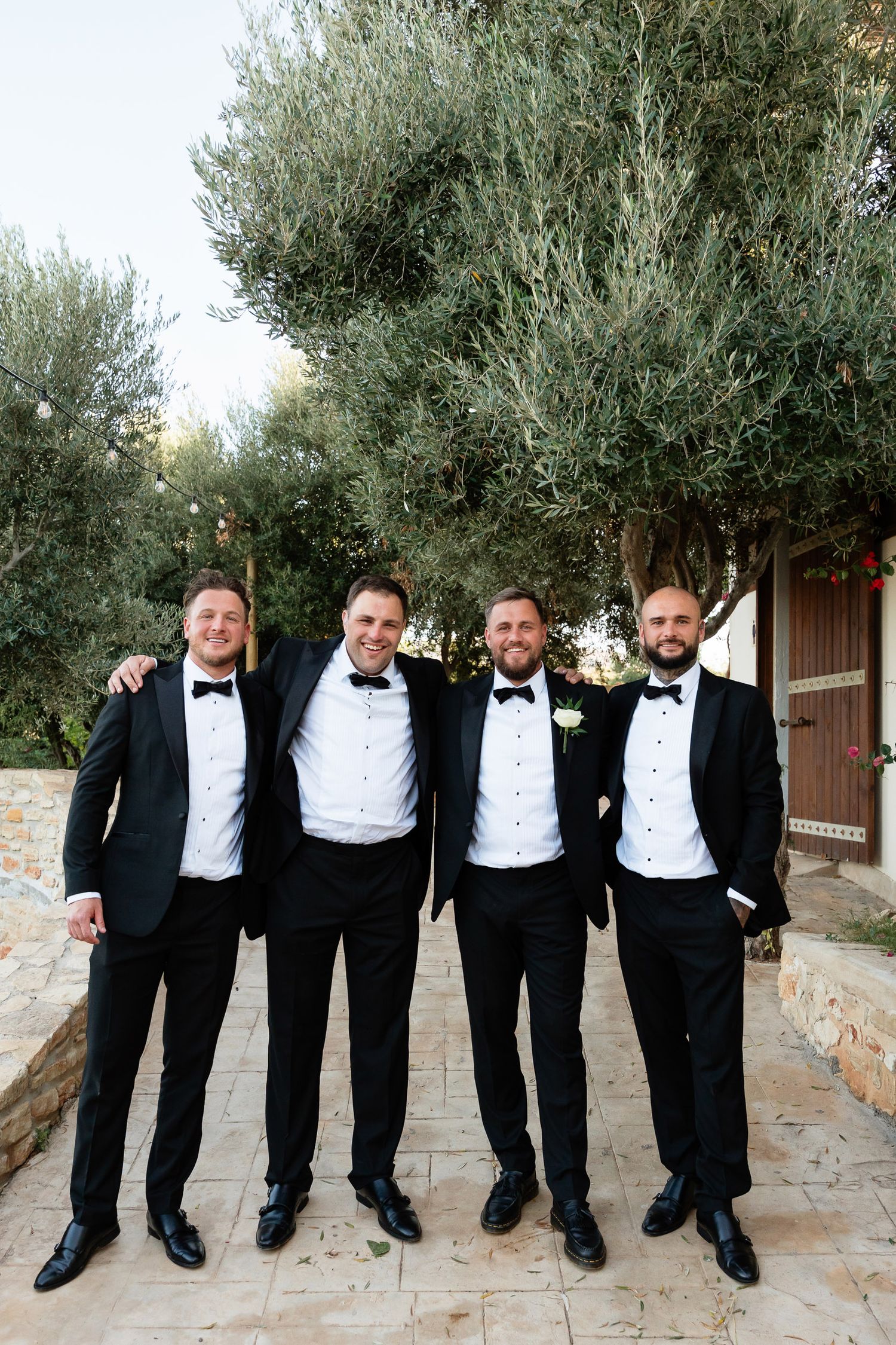 A group of groomsmen in black tuxedos and bowties pose together in a fun sequence of photos on stone steps.