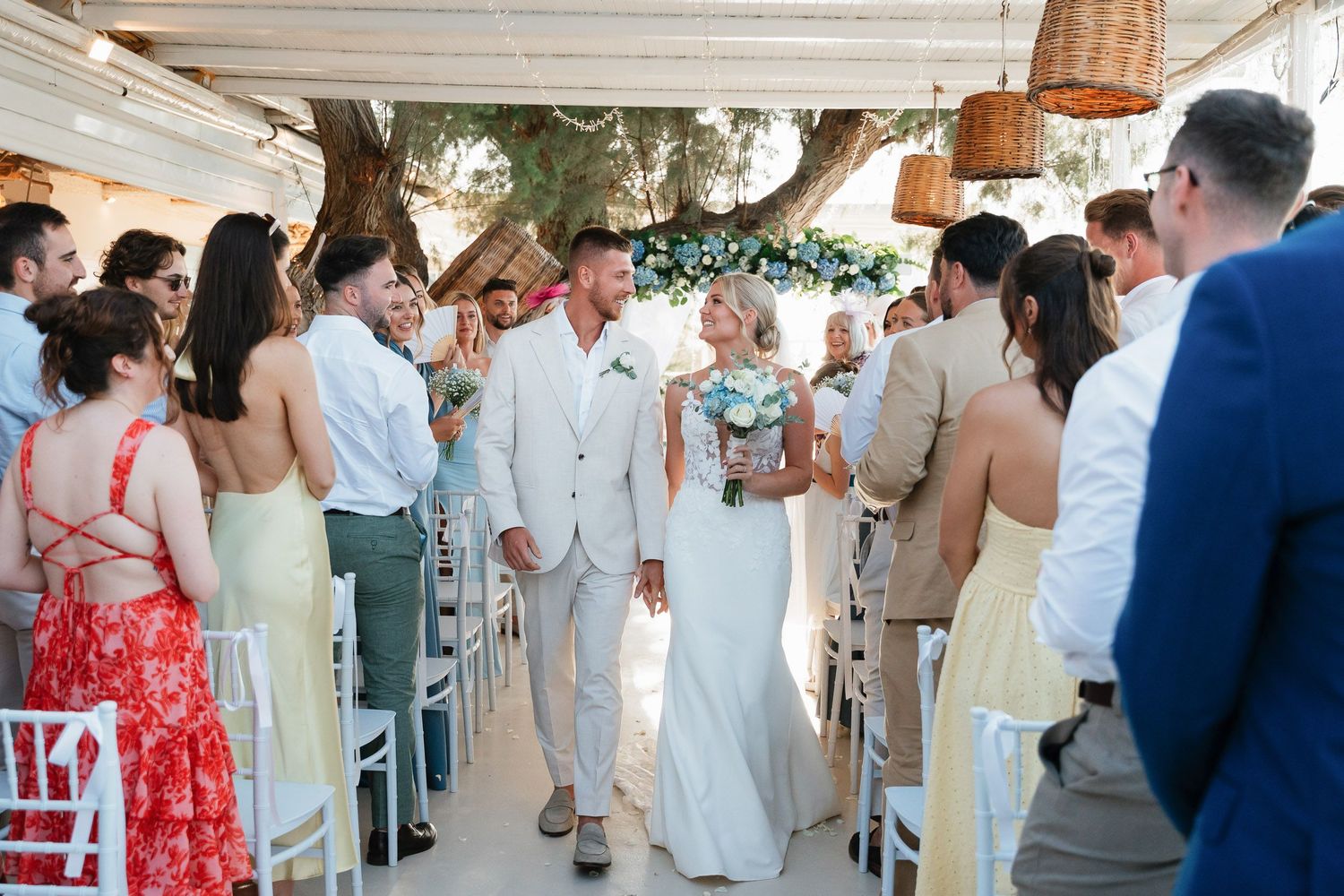 Wedding guests line the aisle as a couple walks down during an outdoor ceremony with hanging basket decor and floral accents.