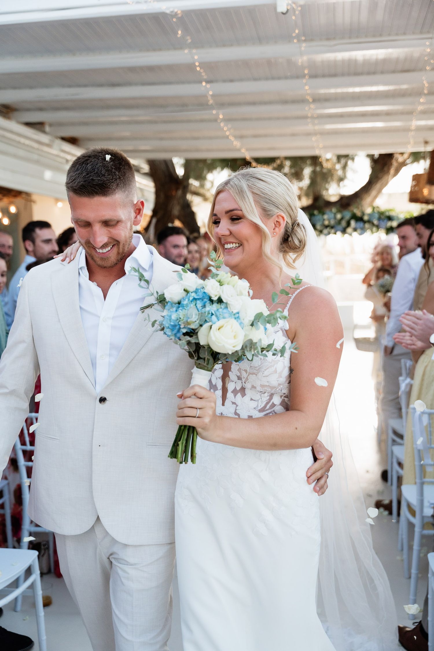 A joyful wedding couple shares intimate moments in white attire with blue and white floral bouquets at their beach ceremony.