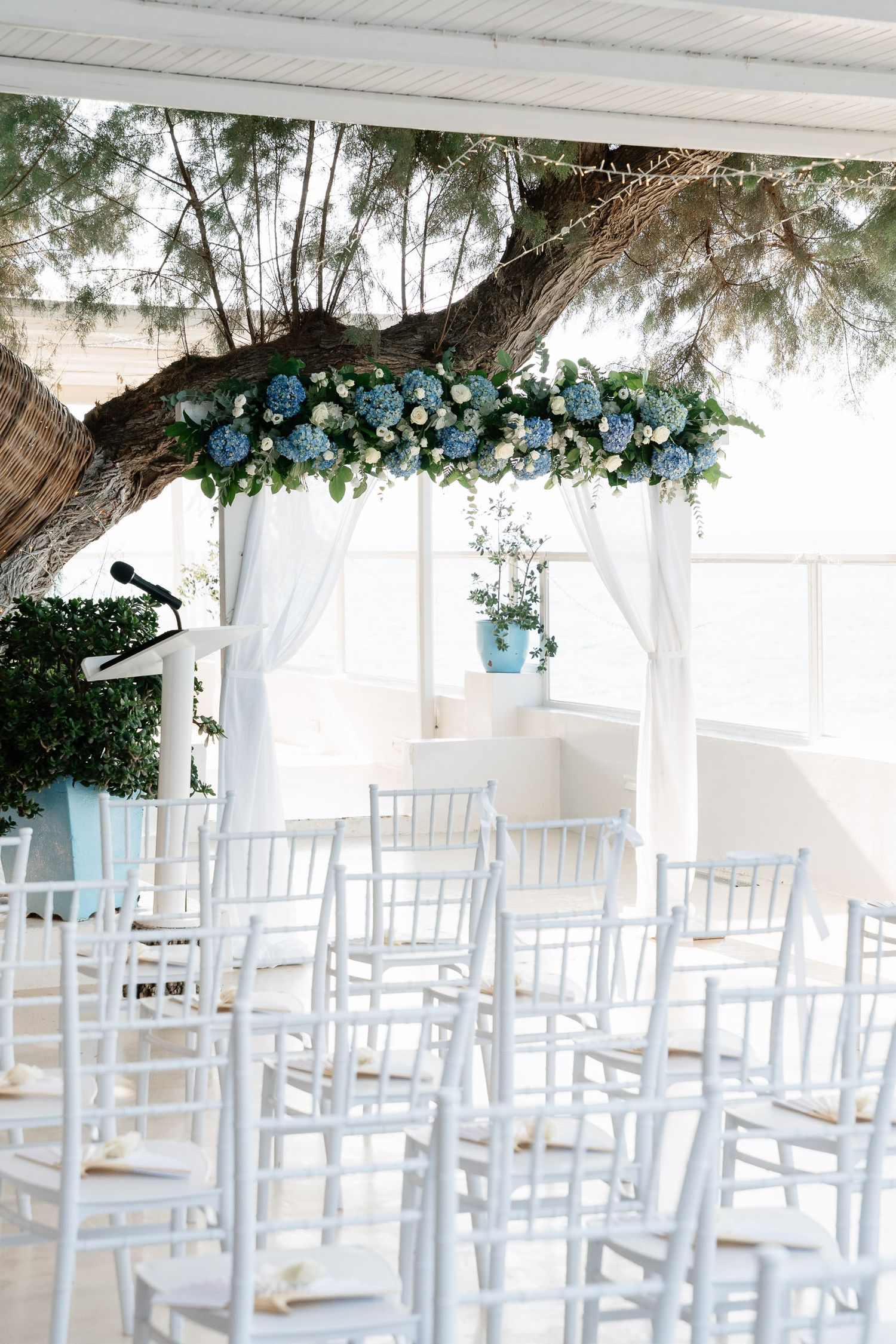 White wedding ceremony setup with elegant chairs and blue floral arch decorations in an airy indoor venue.