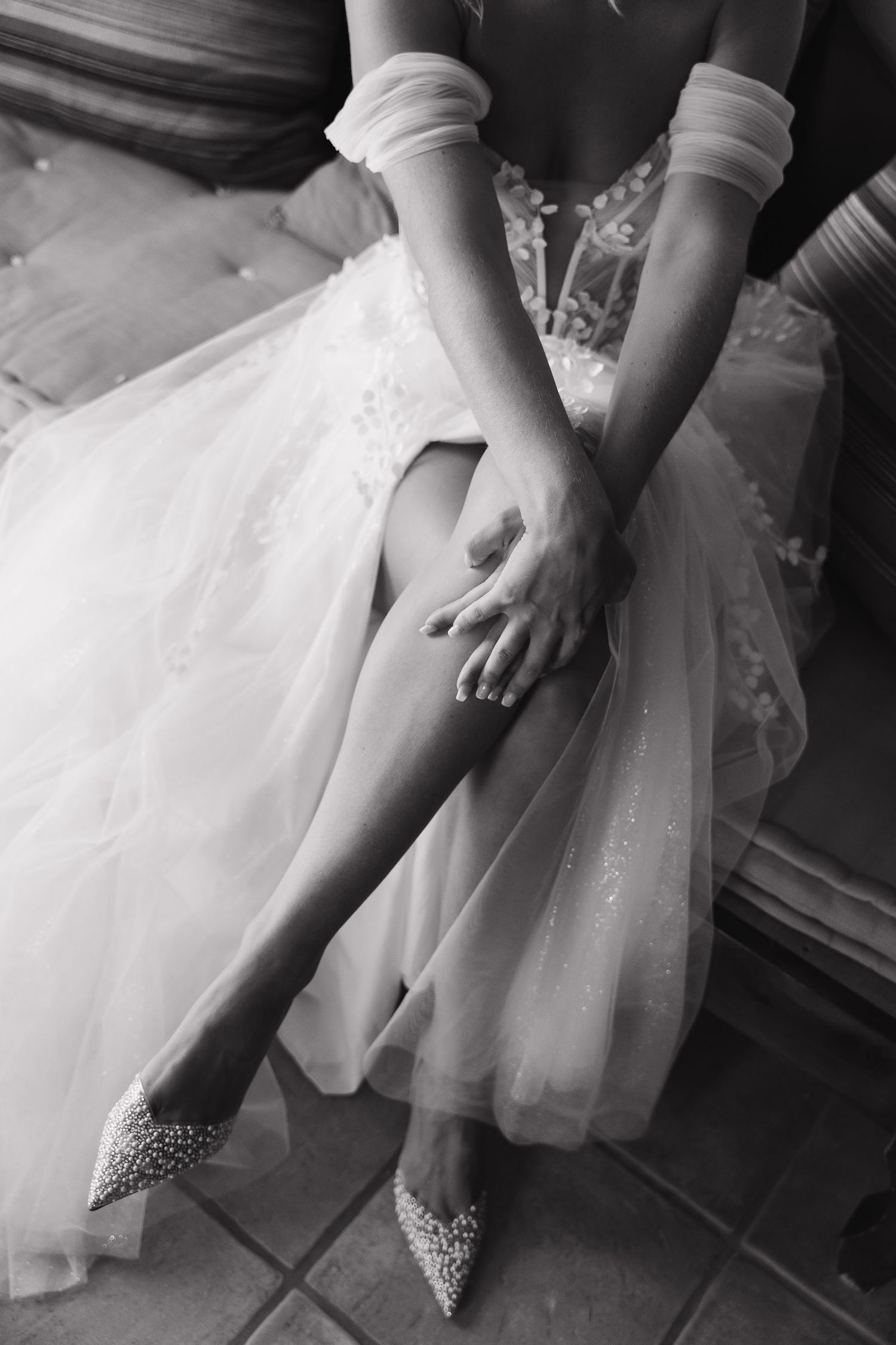 Black and white photo of sparkly bridal shoes and flowing tulle wedding dress fabric with hands resting on knee.