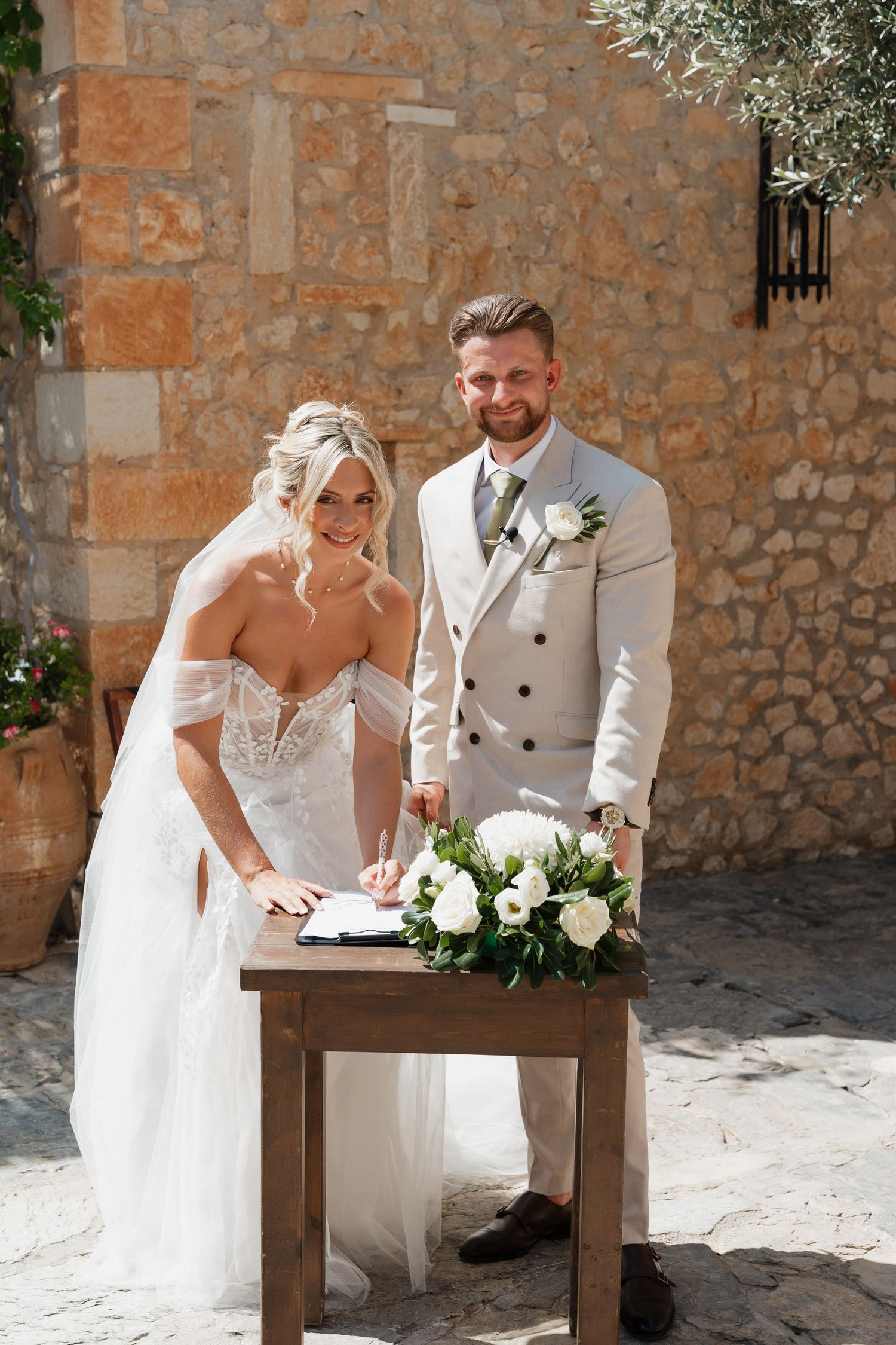 Bride and groom signing wedding documents at rustic stone venue with white floral decorations.