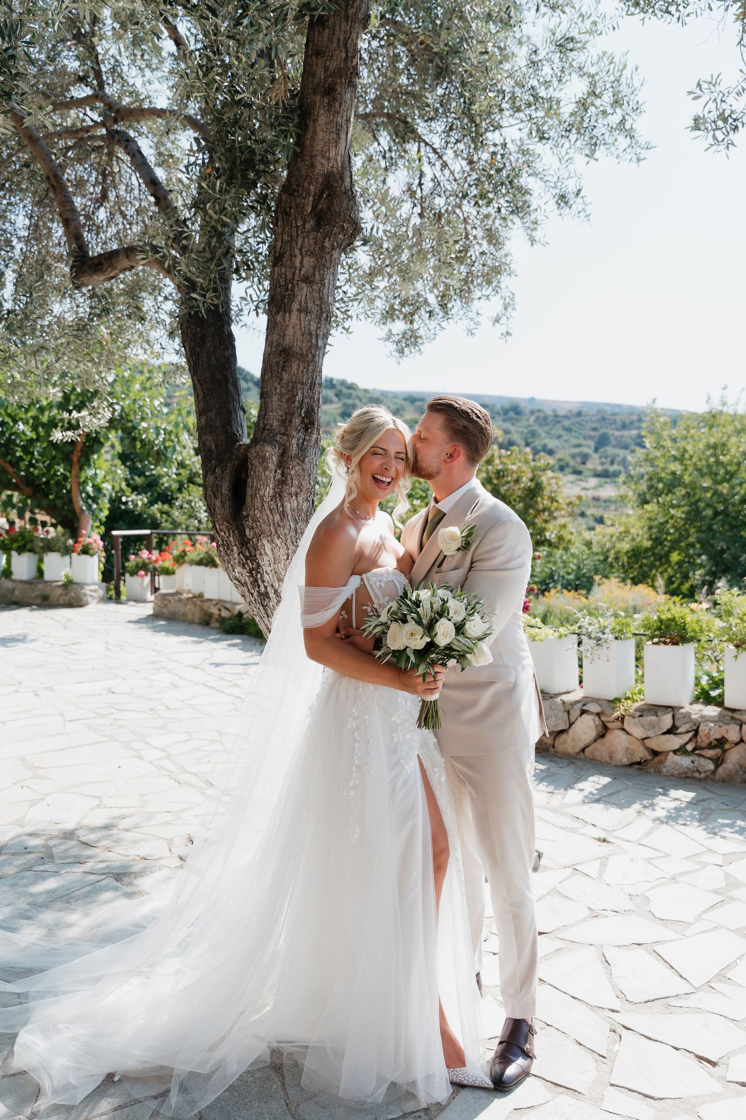 A romantic wedding couple shares intimate moments on a sun-drenched stone terrace surrounded by olive trees.
