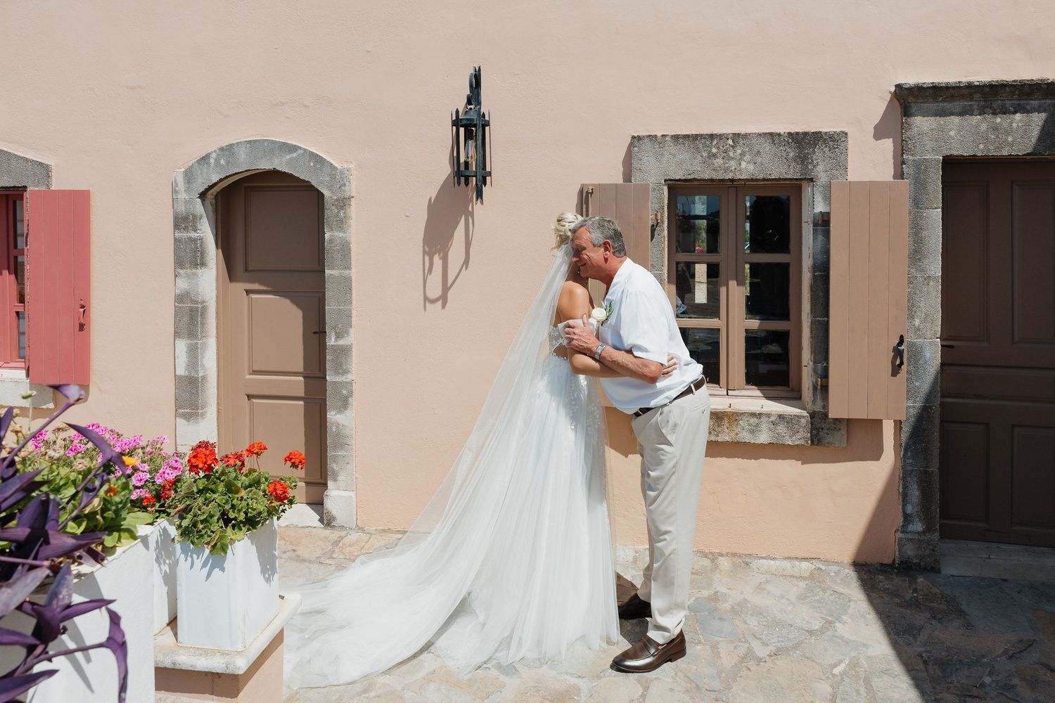 A romantic couple embraces in front of a rustic stone building with pink flowers and wooden shutters in Europe.