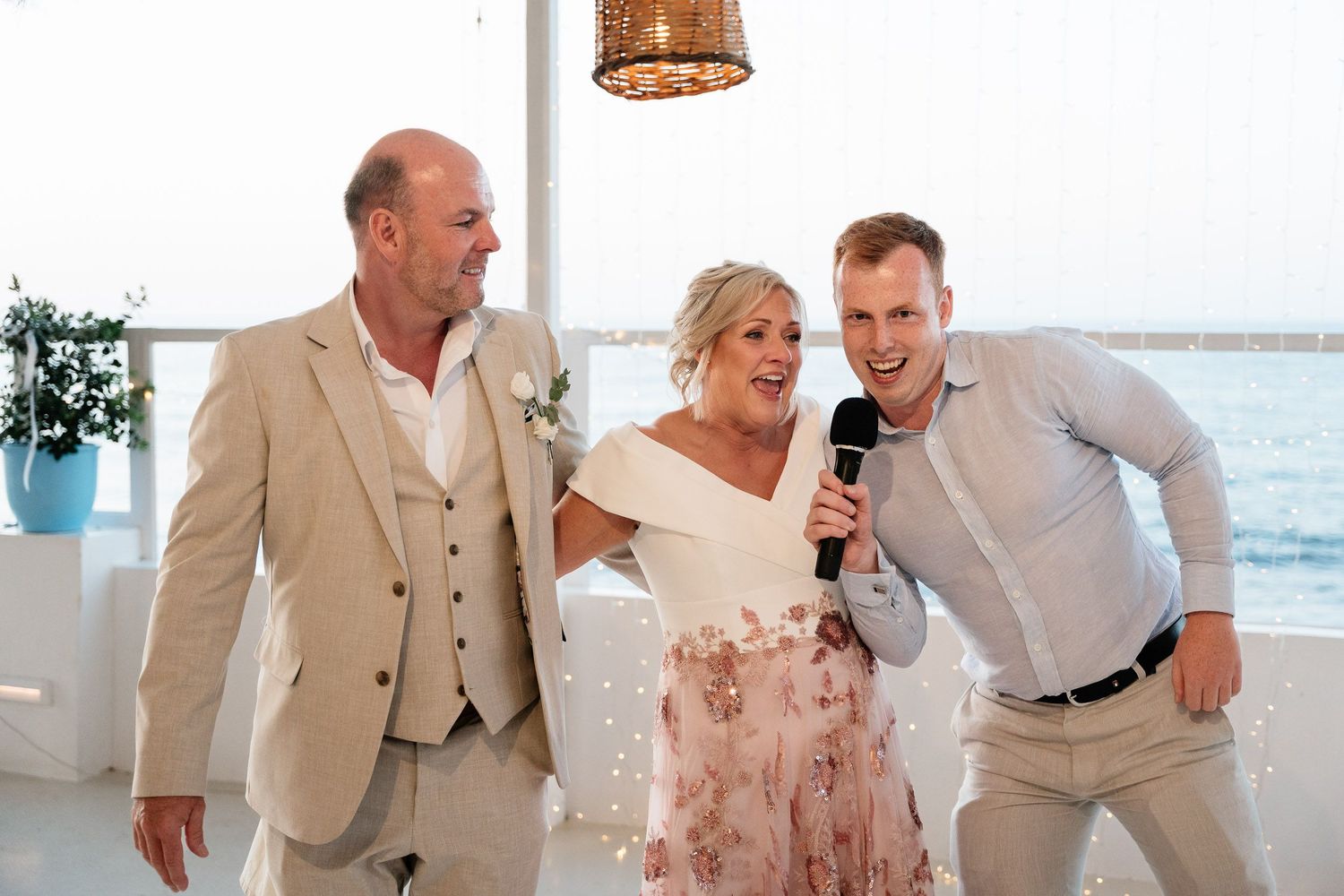 Wedding party members share joyful laughter during a beachside celebration in light-colored formal attire.