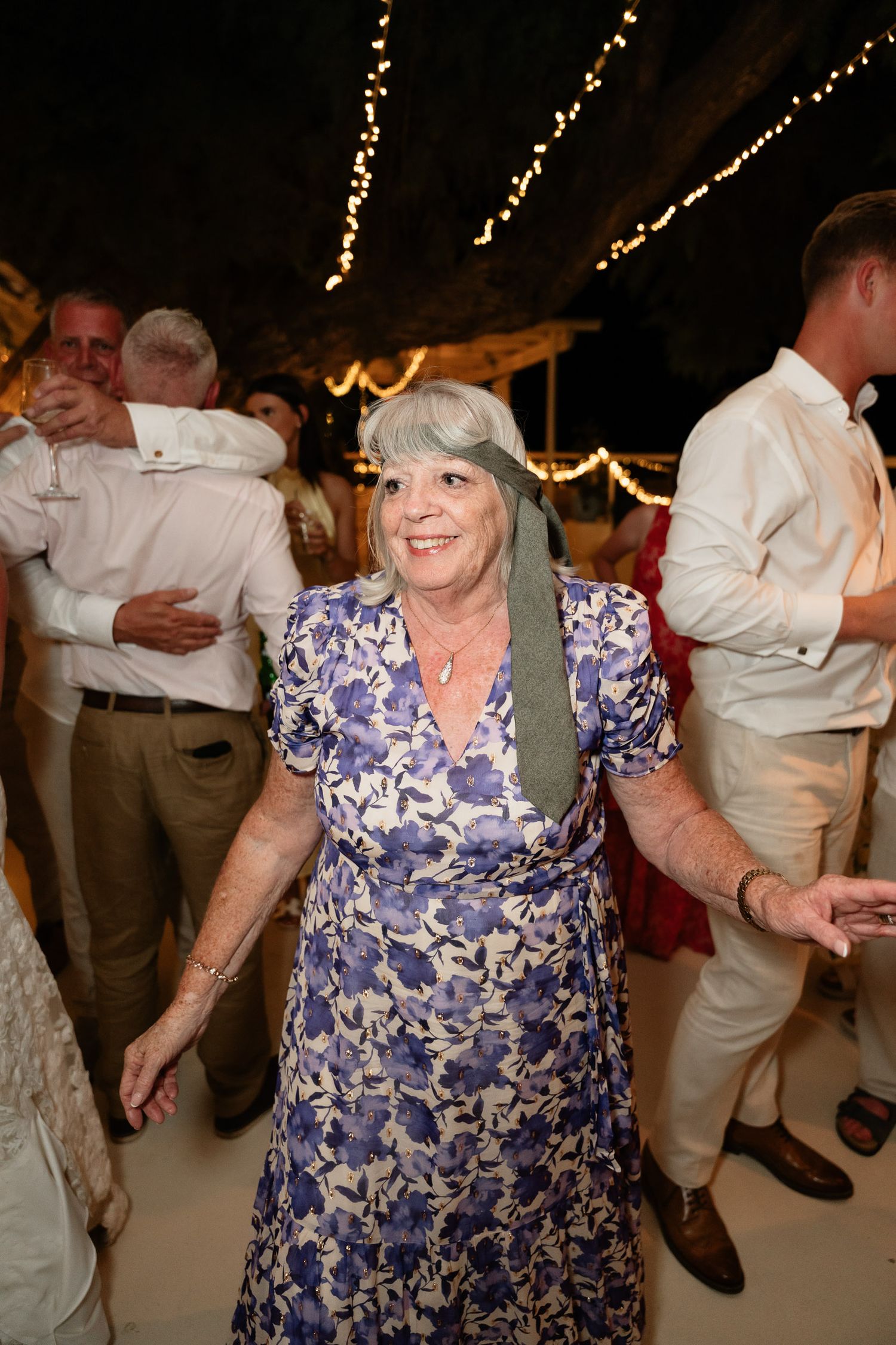 A woman in a blue floral dress dances and smiles at a festive event with string lights overhead.