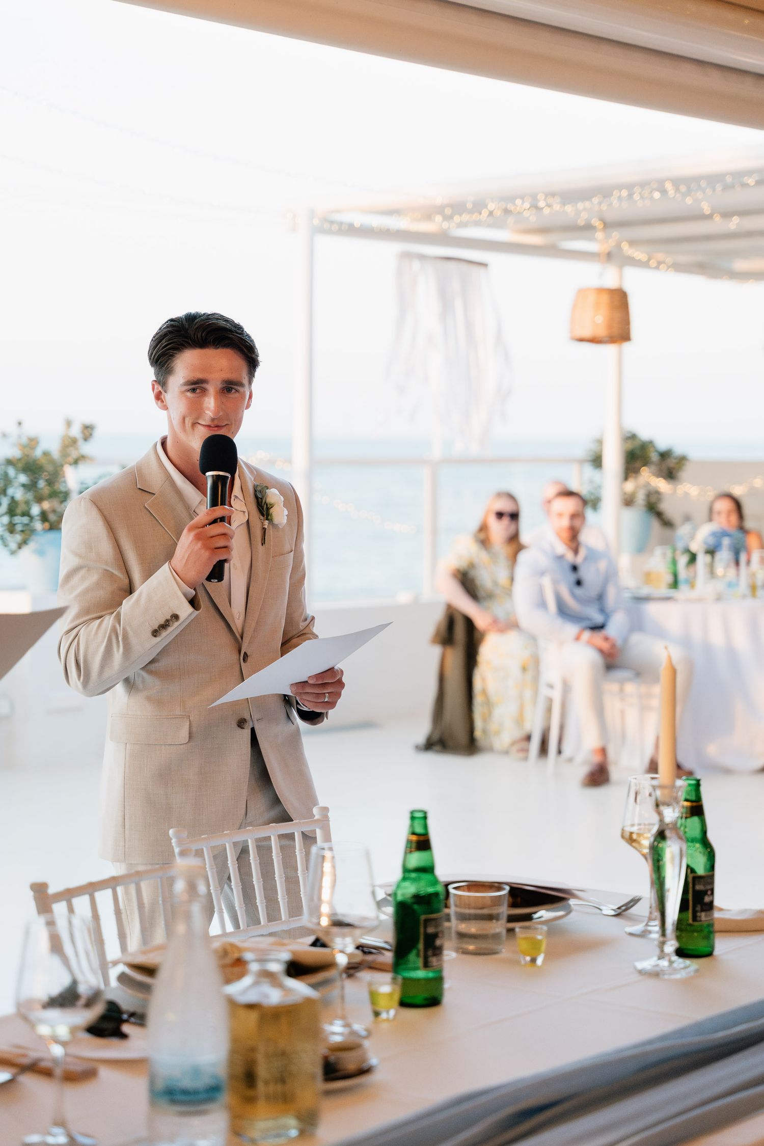 Someone gives a wedding speech at an outdoor beach reception with drinks and table settings visible in the foreground.