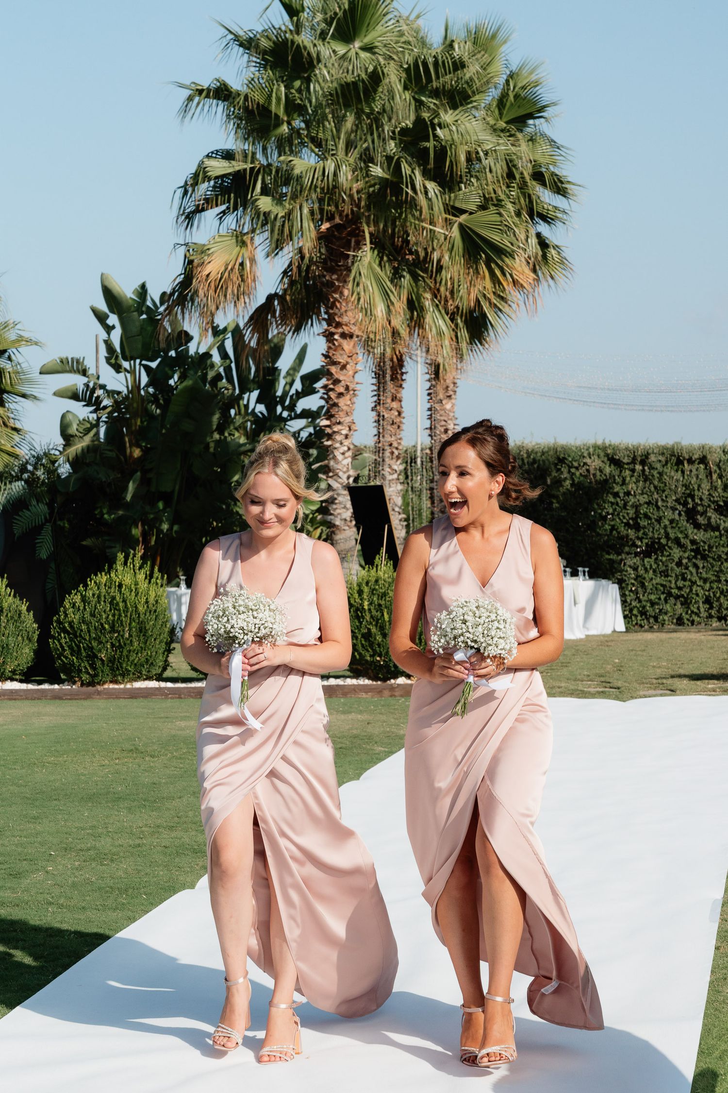 Two bridesmaids in pink dresses and floral tops share a candid moment outdoors with palm trees in the background.