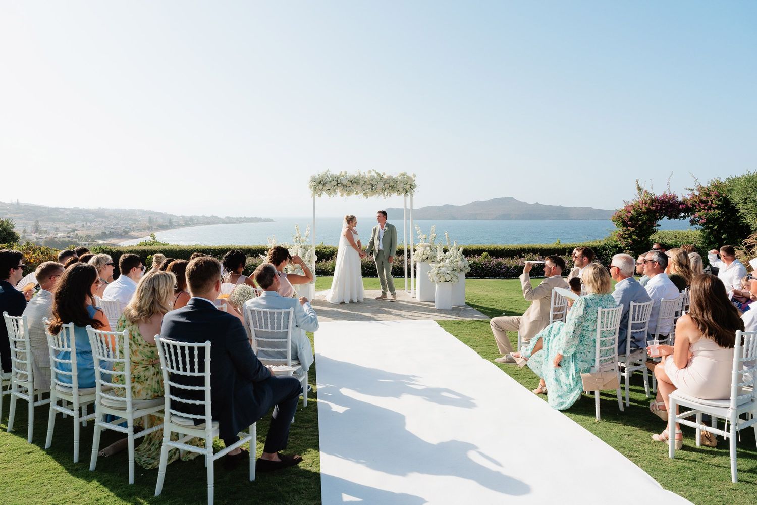 Panoramic view of white chairs set up for outdoor wedding ceremony with ocean and mountains in background.