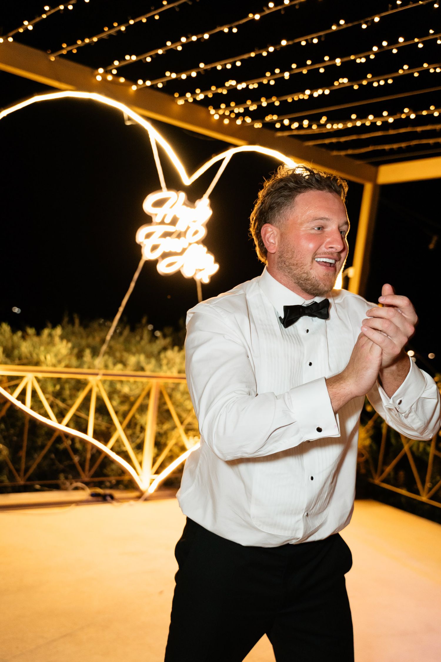 A person in formal attire dances joyfully under twinkling string lights at an outdoor evening celebration.