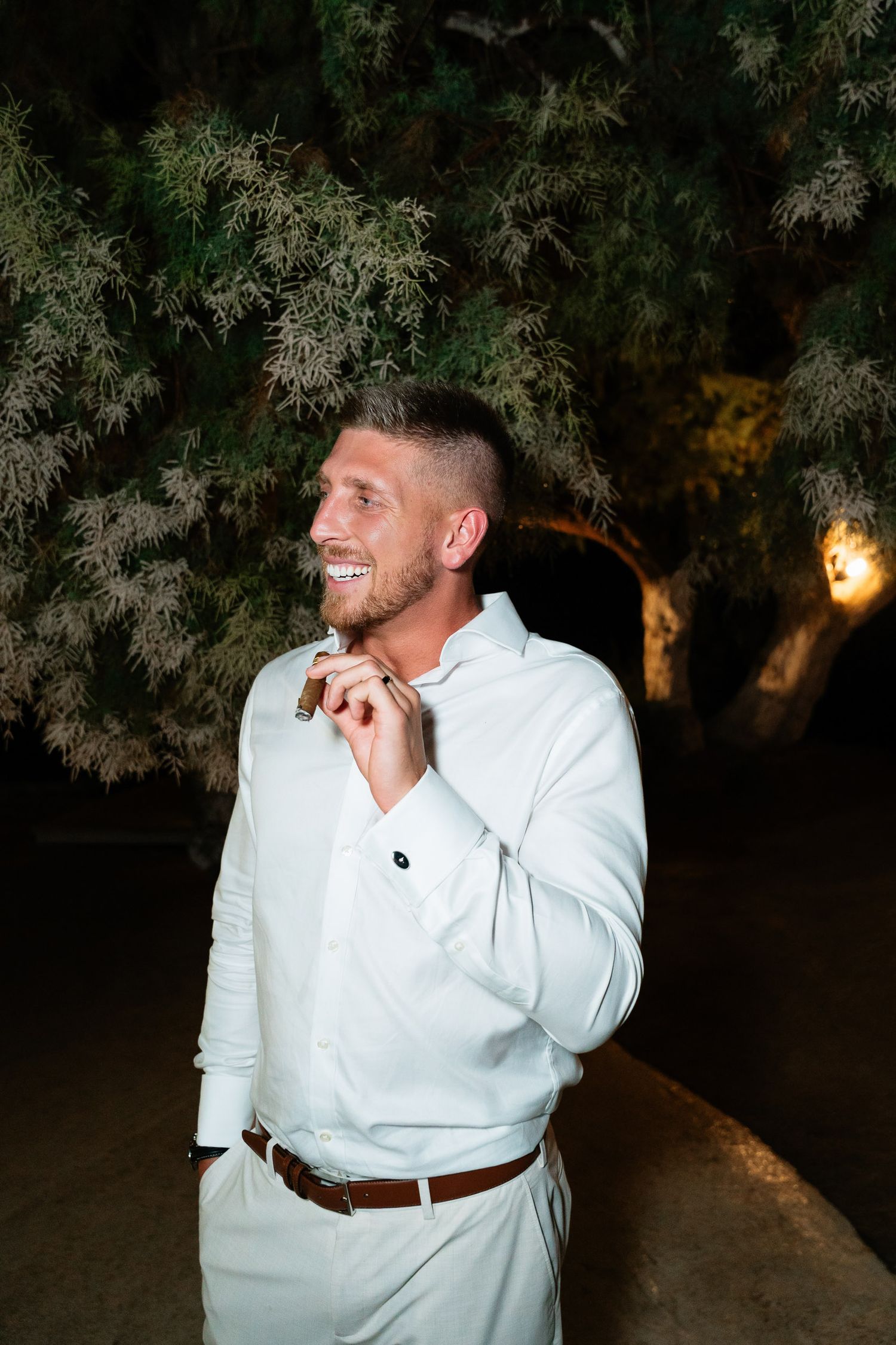 A person in a white button-down shirt and brown belt laughs joyfully while standing outdoors at night.