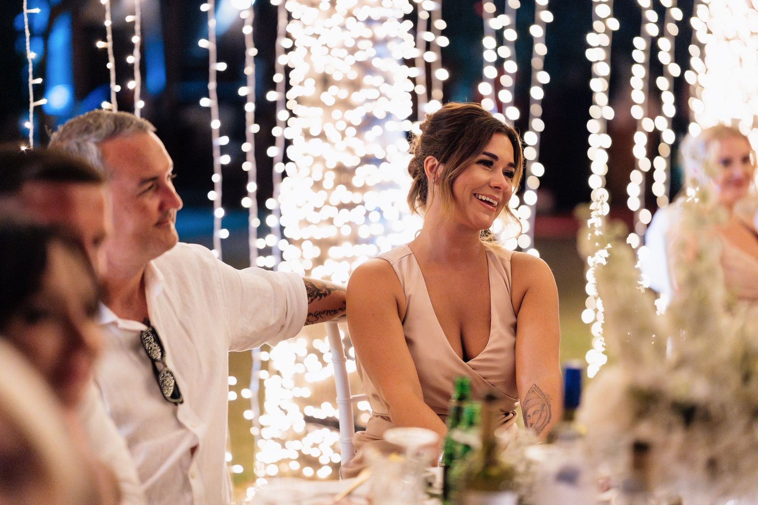 Wedding reception guests seated at elegant tables with hanging string lights and white decor during dinner service.