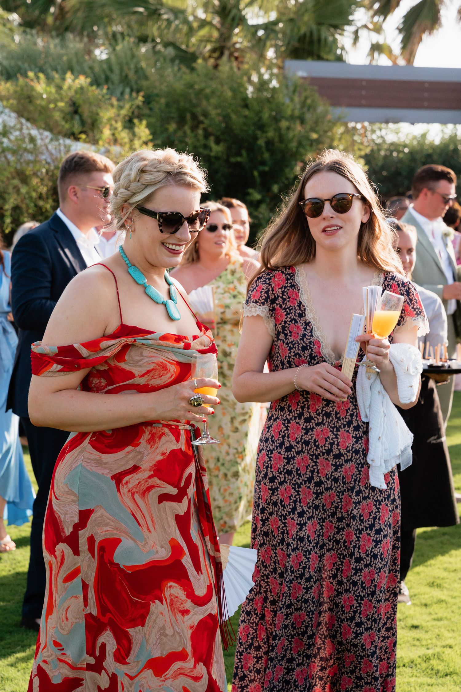 A group enjoying drinks and conversation at an outdoor summer garden party wearing floral and red patterned dresses.