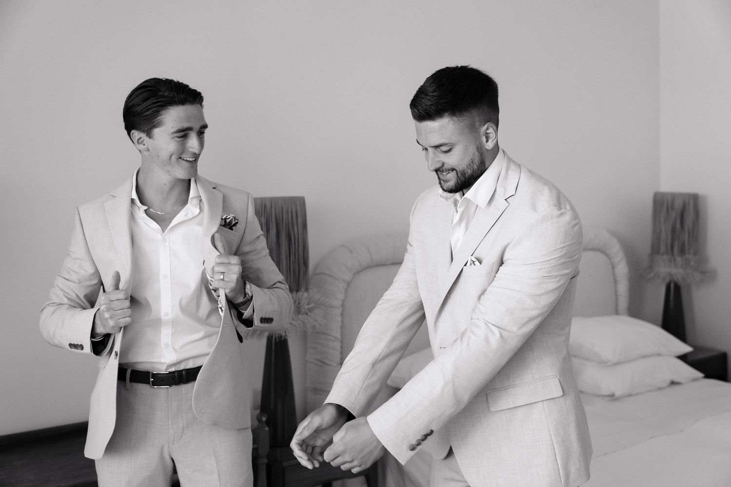Two people in white suits share a lighthearted moment while getting ready in a bedroom setting.