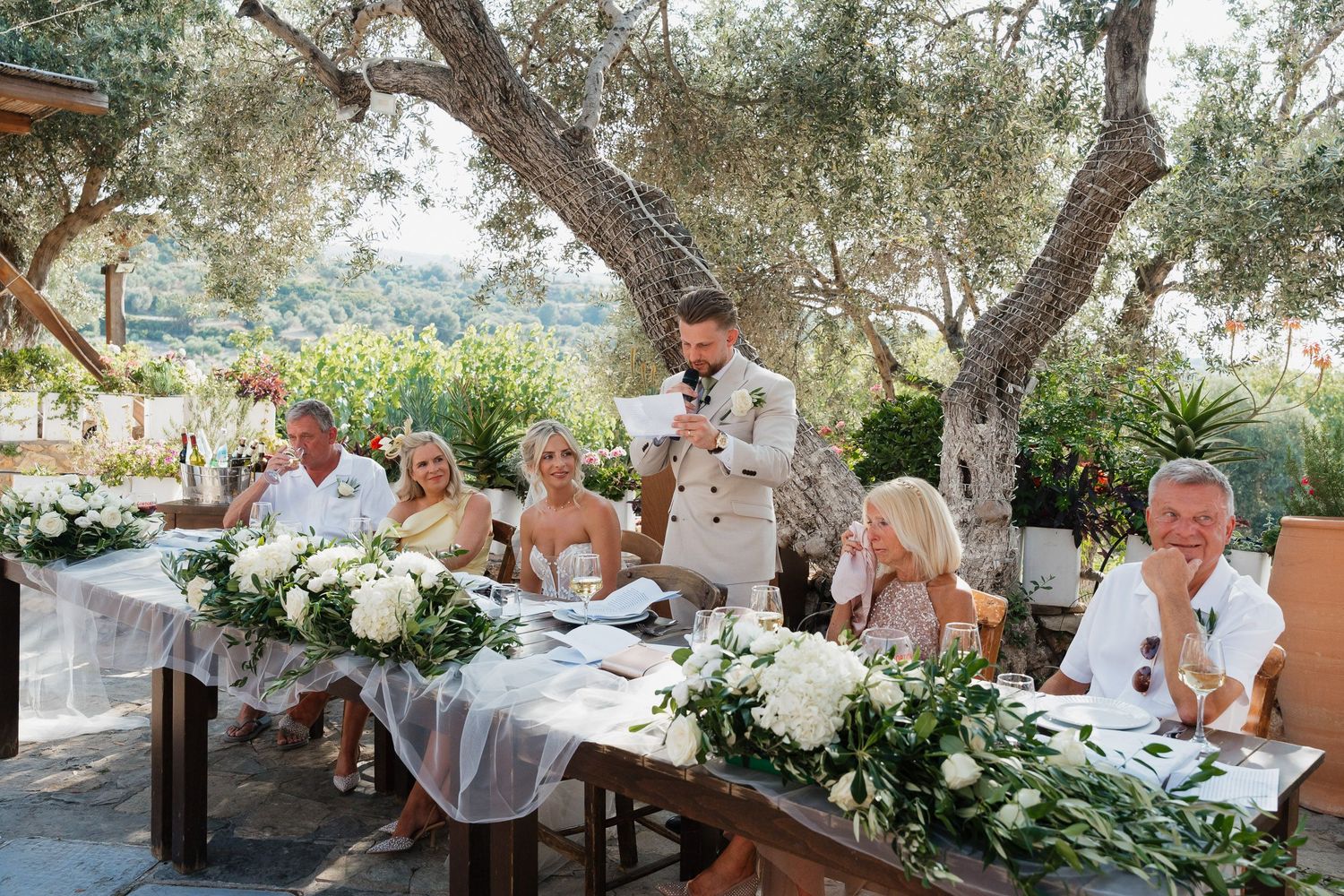 Wedding reception at an outdoor venue with rustic wooden tables decorated with white floral arrangements under olive trees.