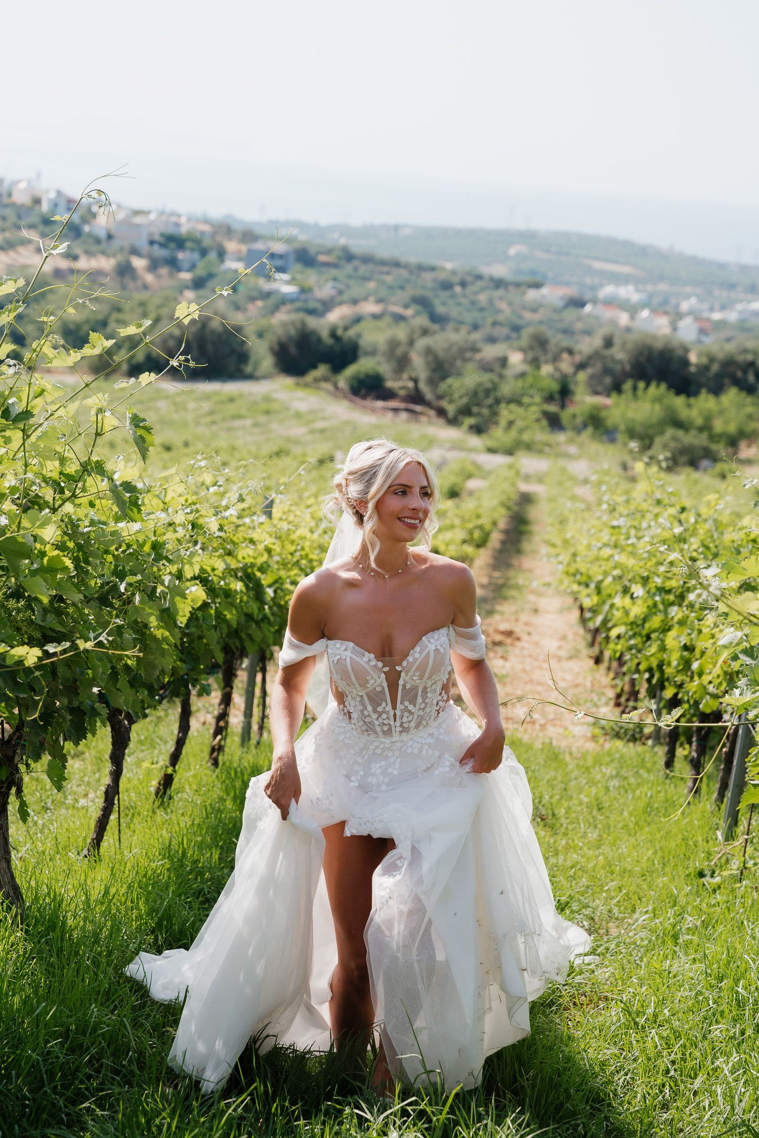 A bride in an elegant off-shoulder wedding gown walks through a scenic vineyard on a hillside during a sunny day.