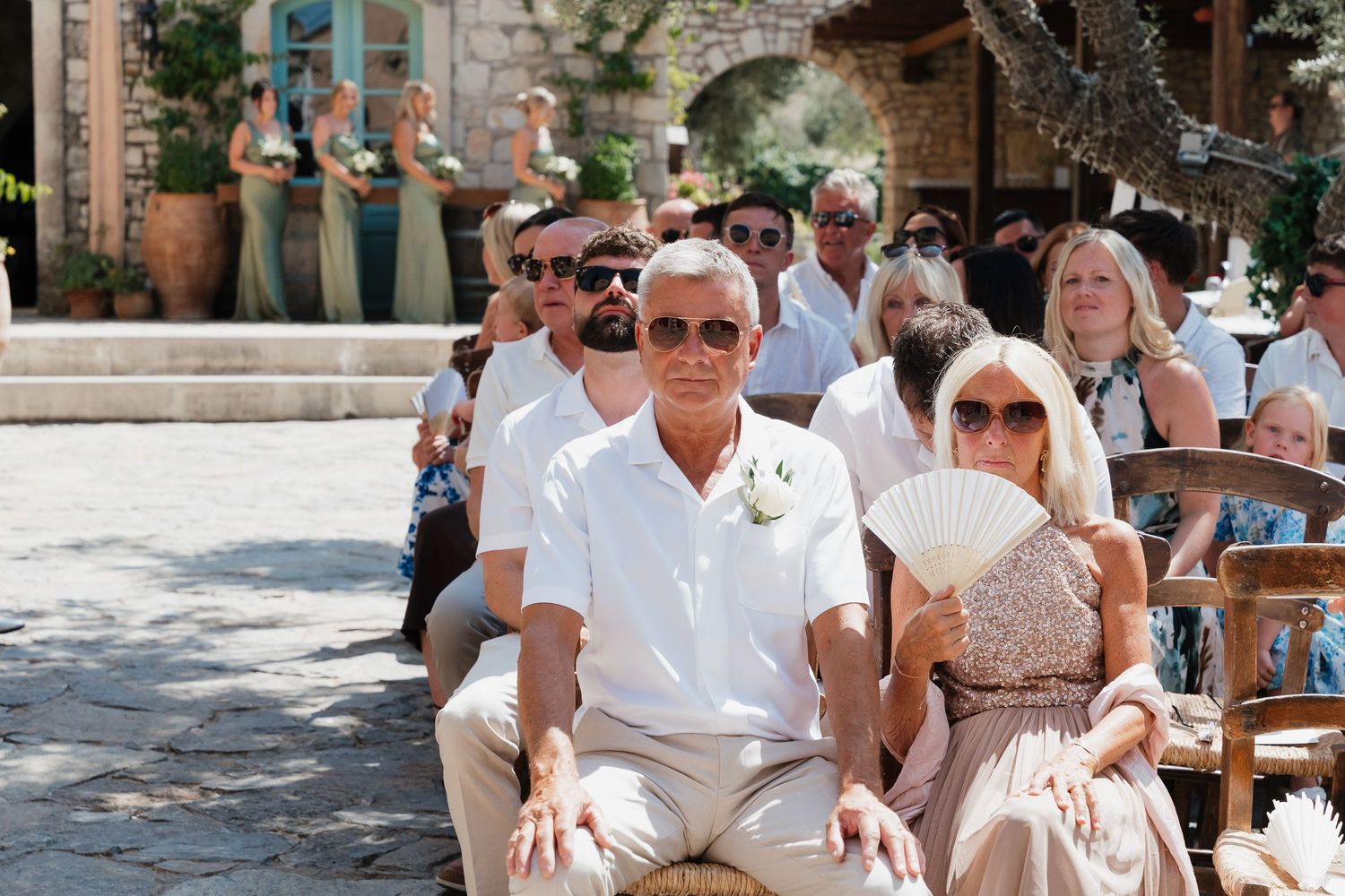 Wedding guests in white and light colored attire sit in rows at an outdoor ceremony venue on a sunny day.