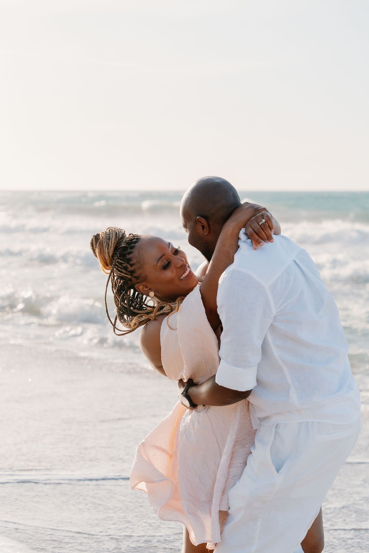 A couple in white outfits shares an intimate moment on a beach at sunset.