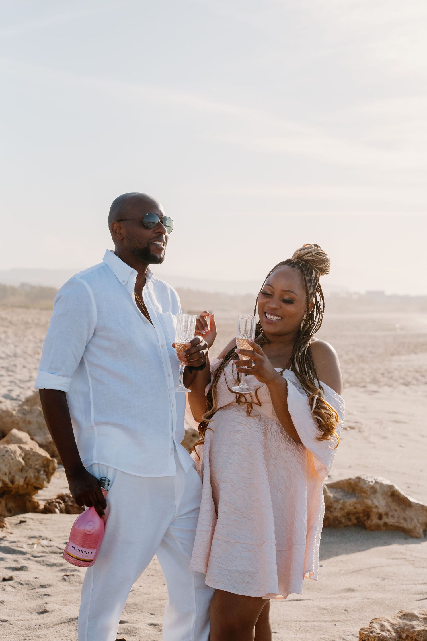 A couple in white outfits enjoying drinks together on a beach at sunset.