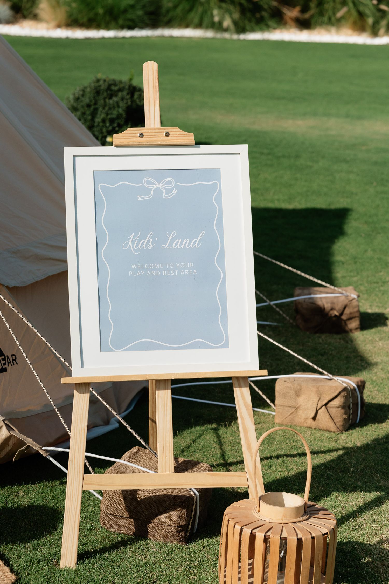 A wooden easel displays a light blue framed welcome sign on a grassy lawn with wooden crates placed nearby.