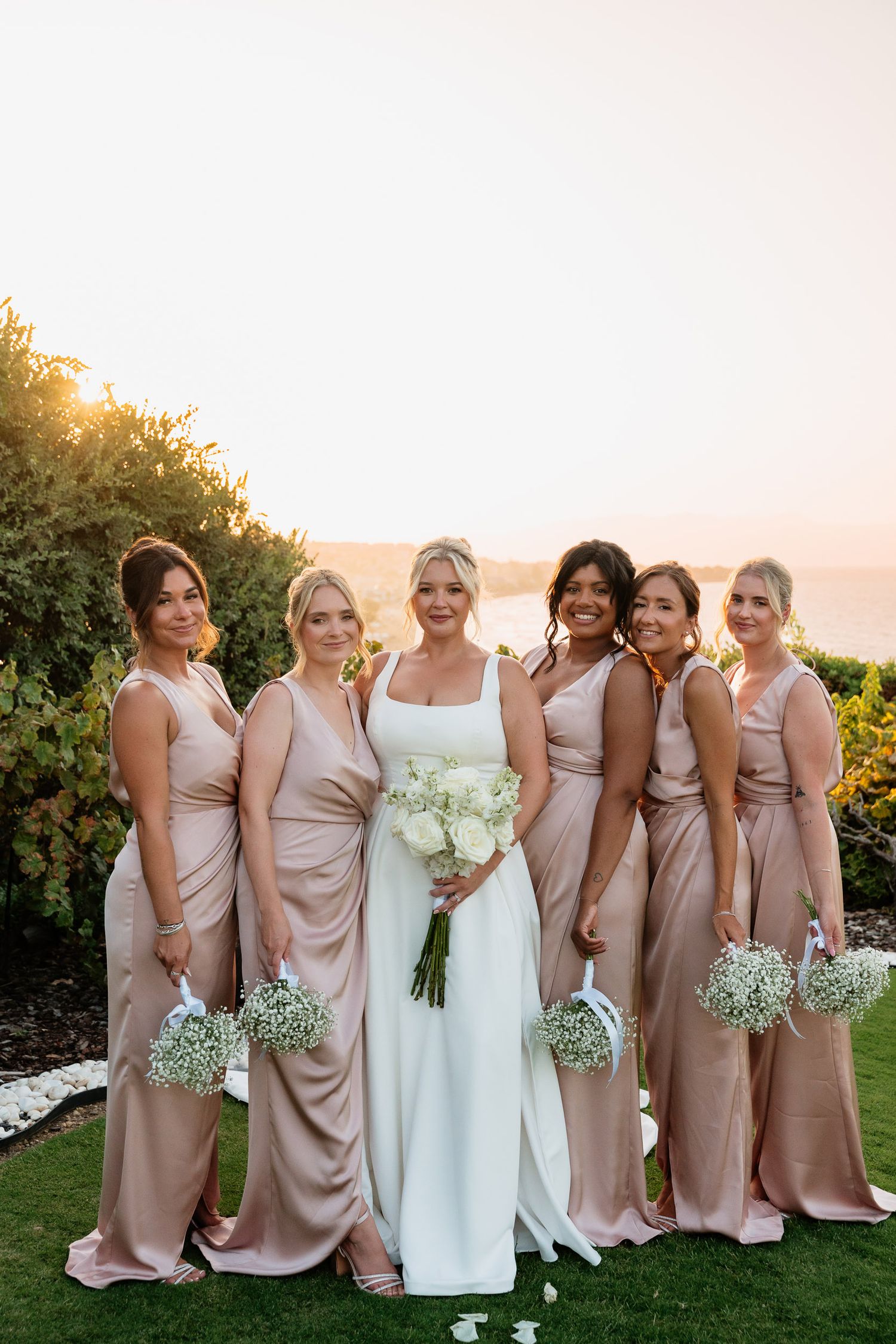 A bridal party in blush pink dresses poses together on a grassy lawn at sunset.