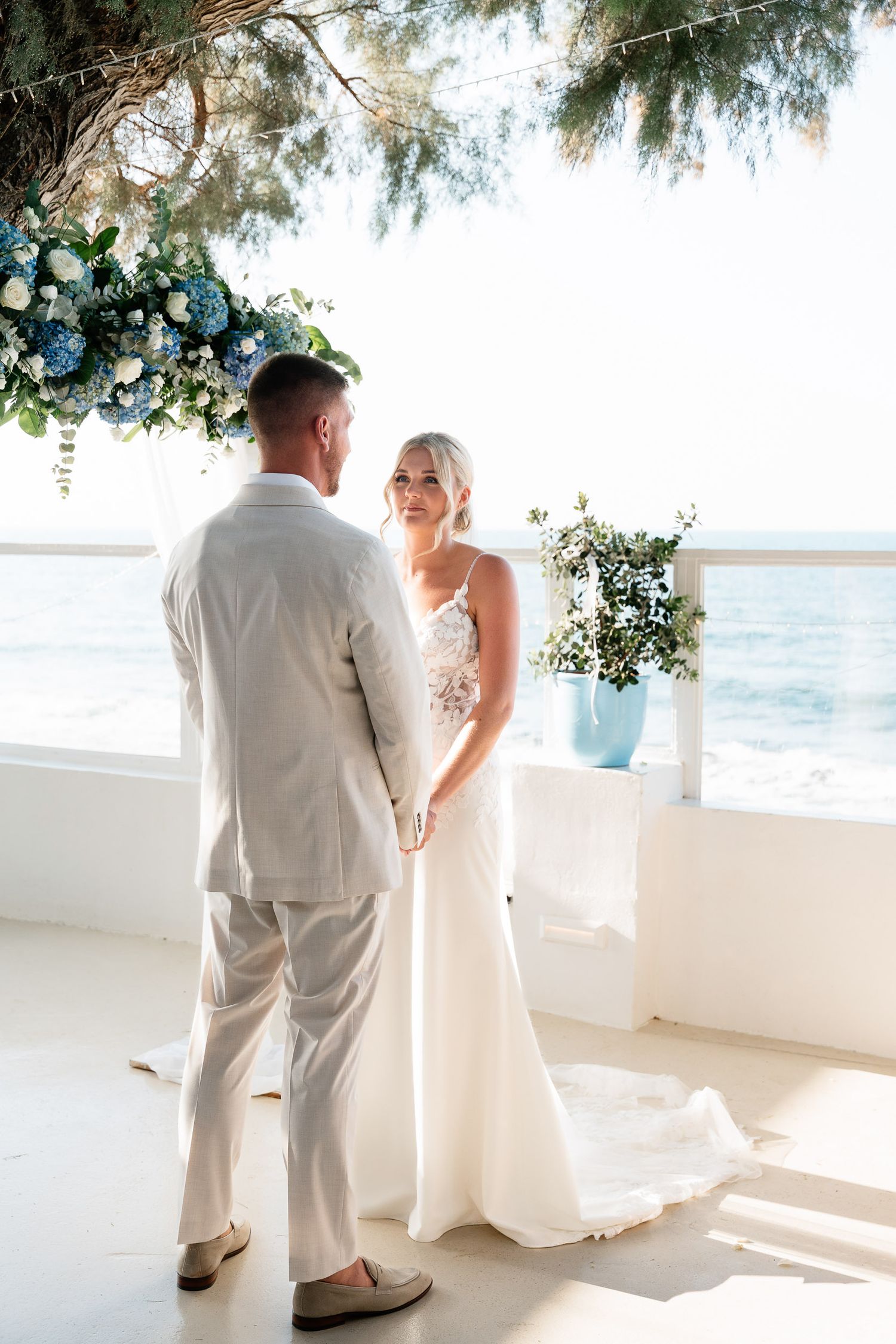 A couple in white attire shares an intimate moment on a sunny balcony overlooking the ocean.