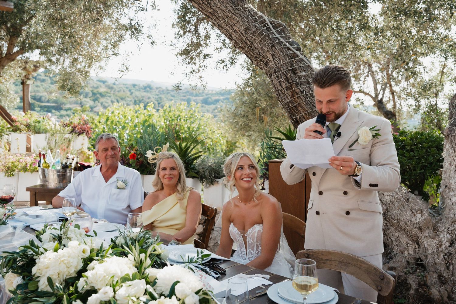 Wedding guests seated at an elegant outdoor reception table under an olive tree during a daytime celebration.