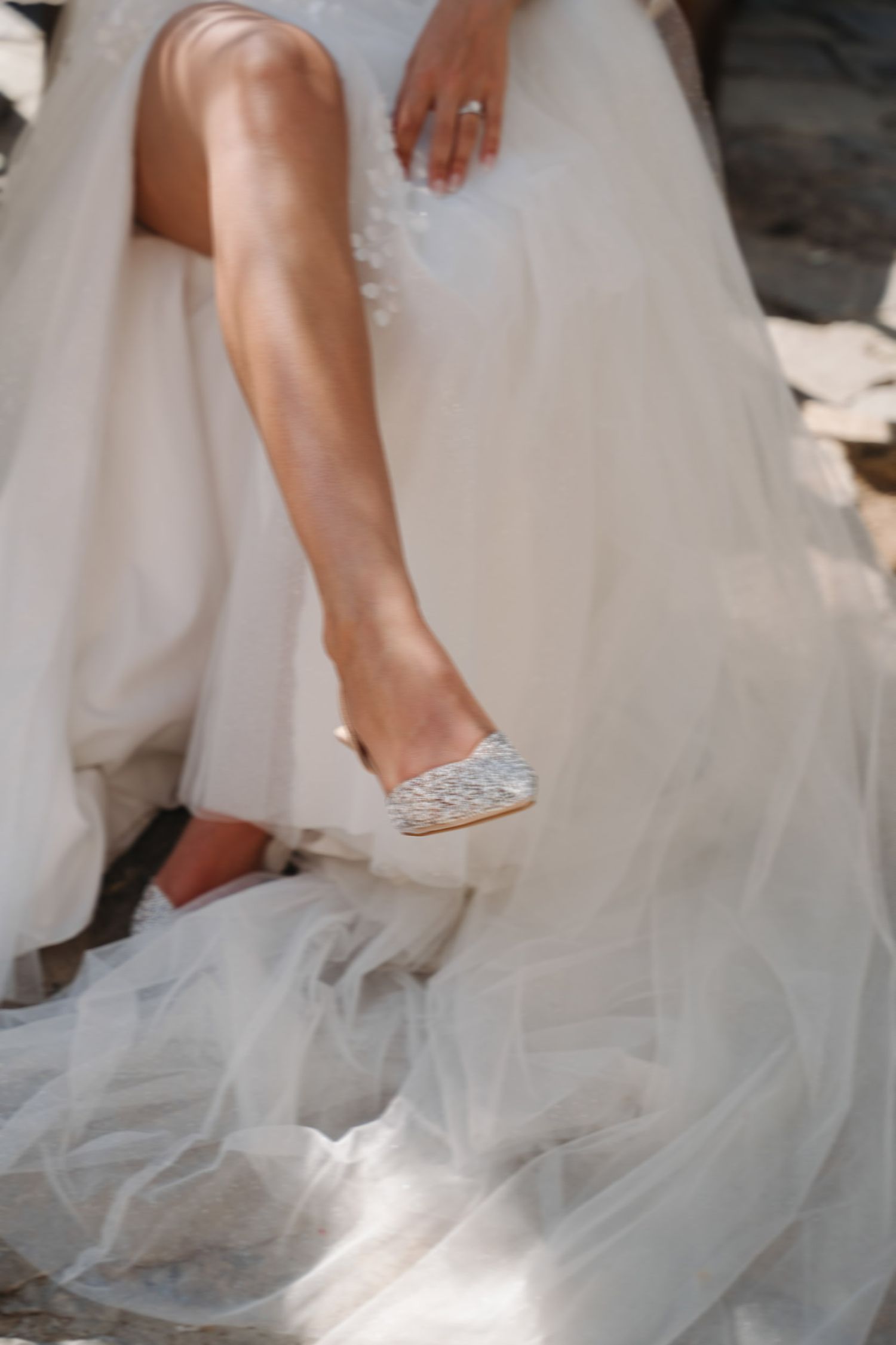 Close-up views of elegant bridal shoes with crystal embellishments peeking beneath layers of flowing white tulle gown.