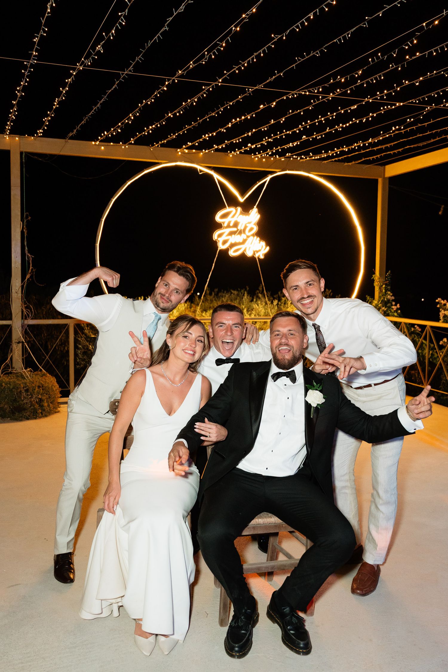 Wedding party members dance and pose together on a white dance floor under twinkling string lights at an evening reception.