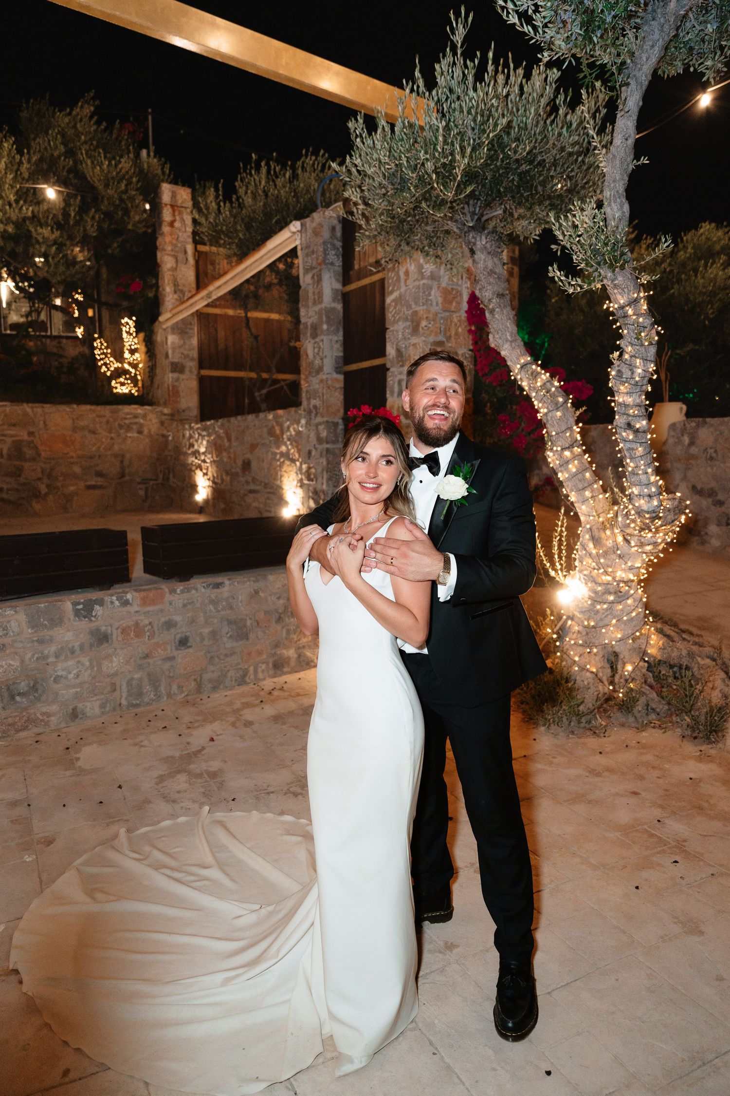 Couple dancing together in snow-covered outdoor venue with illuminated trees and stone walls in the background.