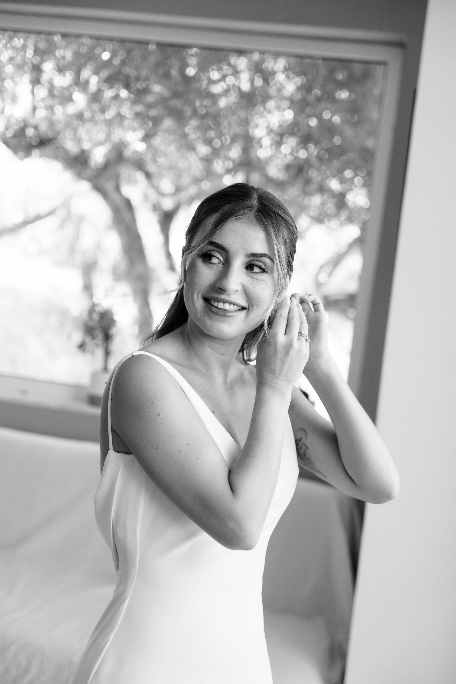 Black and white portrait of someone in a white sleeveless dress putting on earrings near a bright window.