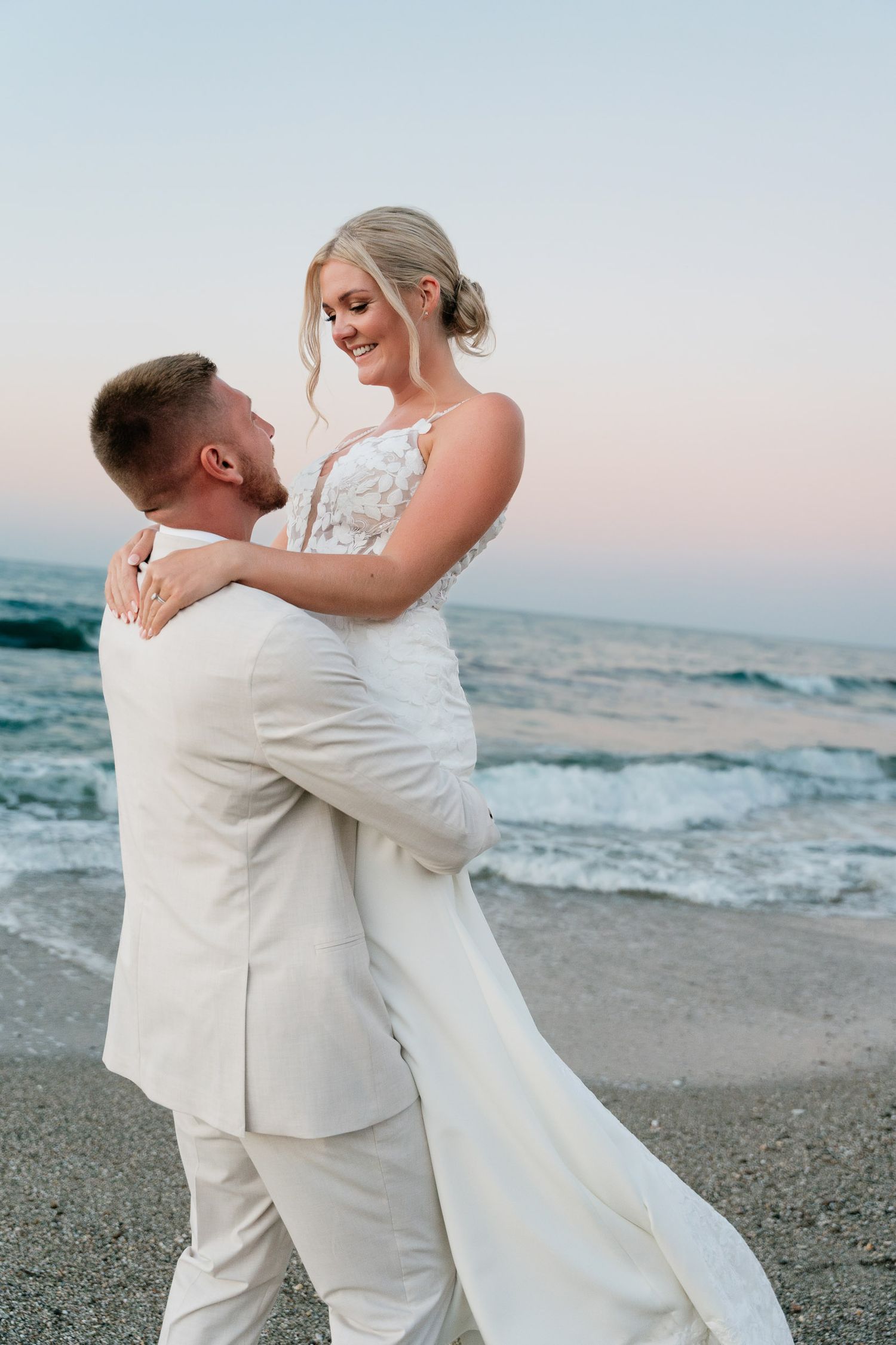 A couple in white wedding attire embraces romantically on a beach at sunset with ocean waves in the background.