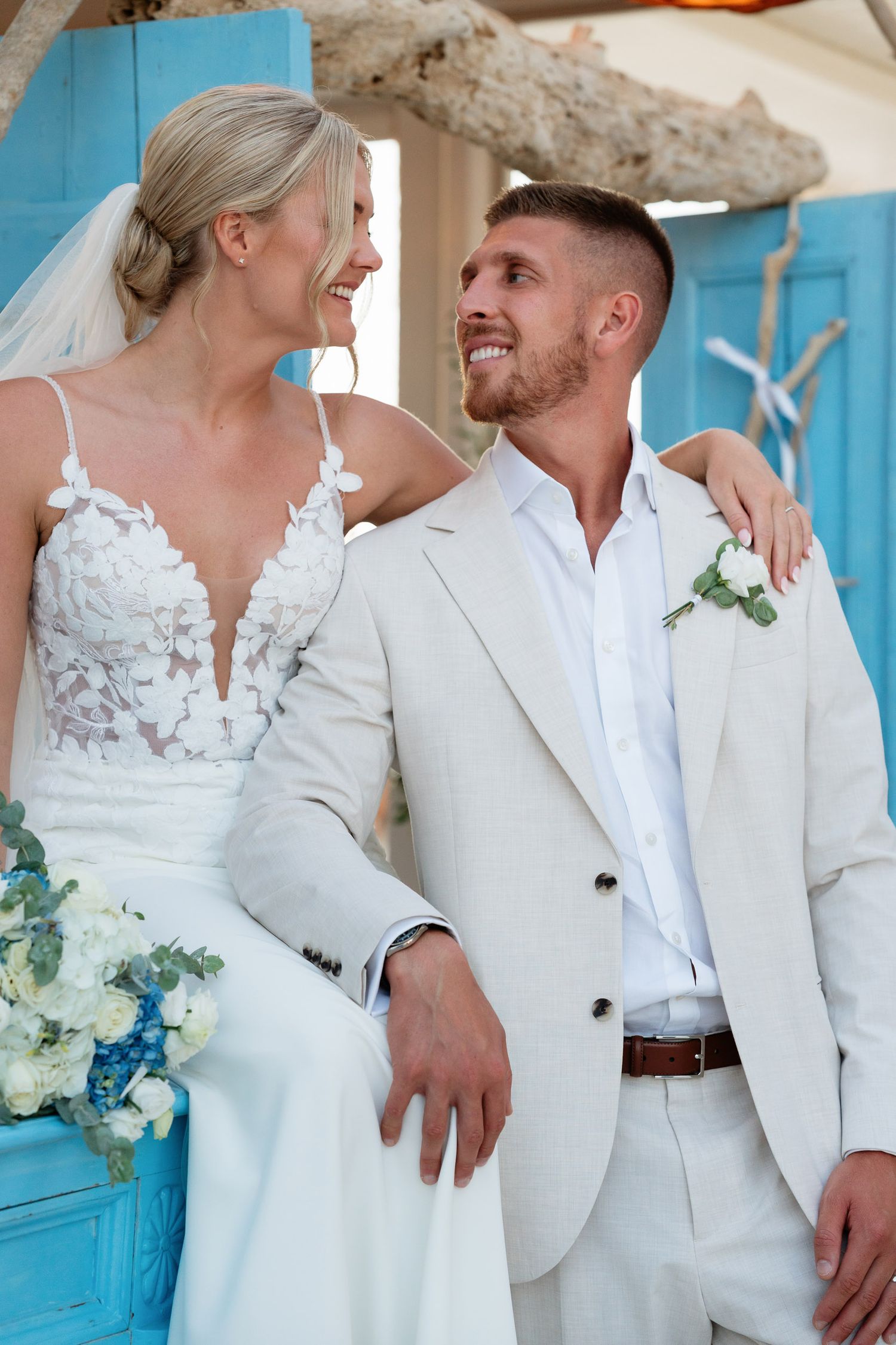 A newlywed couple in white attire share a romantic moment against a turquoise backdrop during their beach wedding.