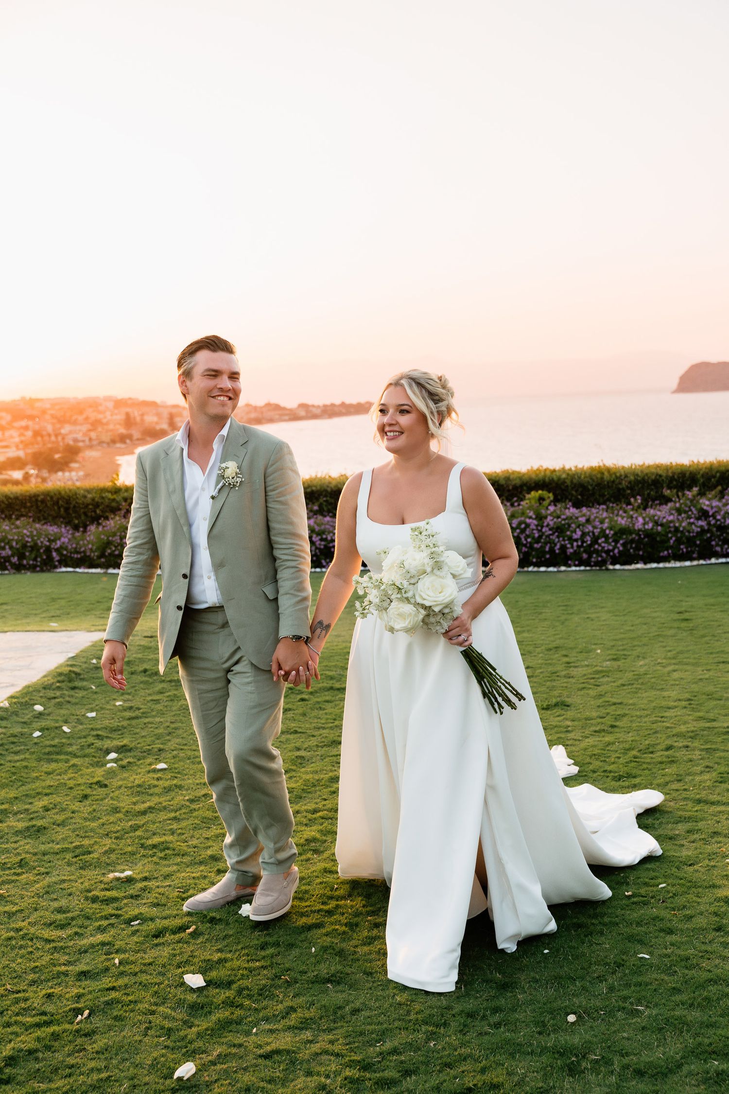 A couple in wedding attire walks on a manicured lawn at sunset with an ocean view in the background.