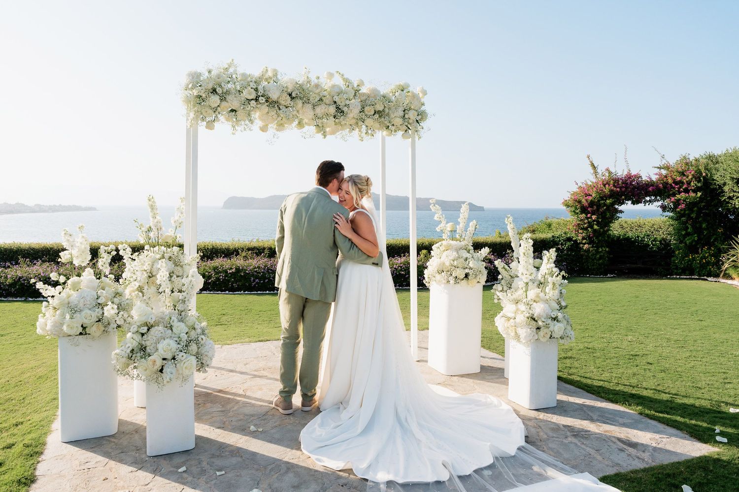 A wedding ceremony setup on a lush green lawn with white floral arch and decorative pillars against an ocean backdrop.