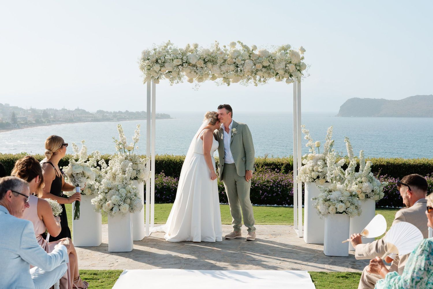 A romantic seaside wedding ceremony with floral arch decorations and guests seated on white chairs overlooking the ocean.