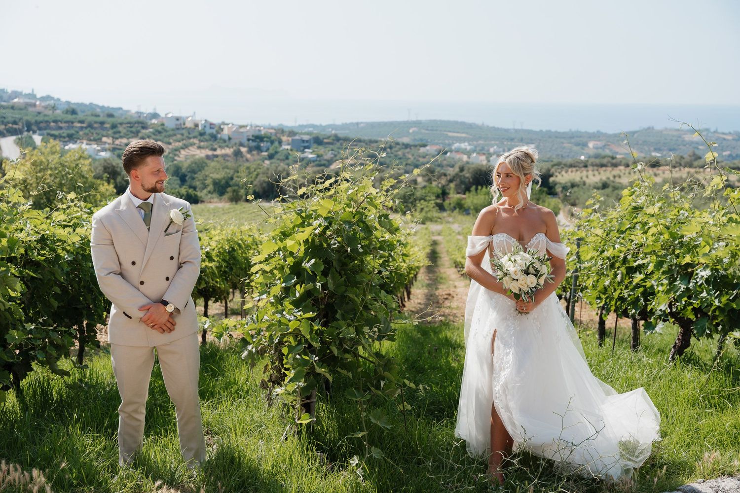 A couple stands in a sun-drenched vineyard on their wedding day with rolling hills and greenery in the background.