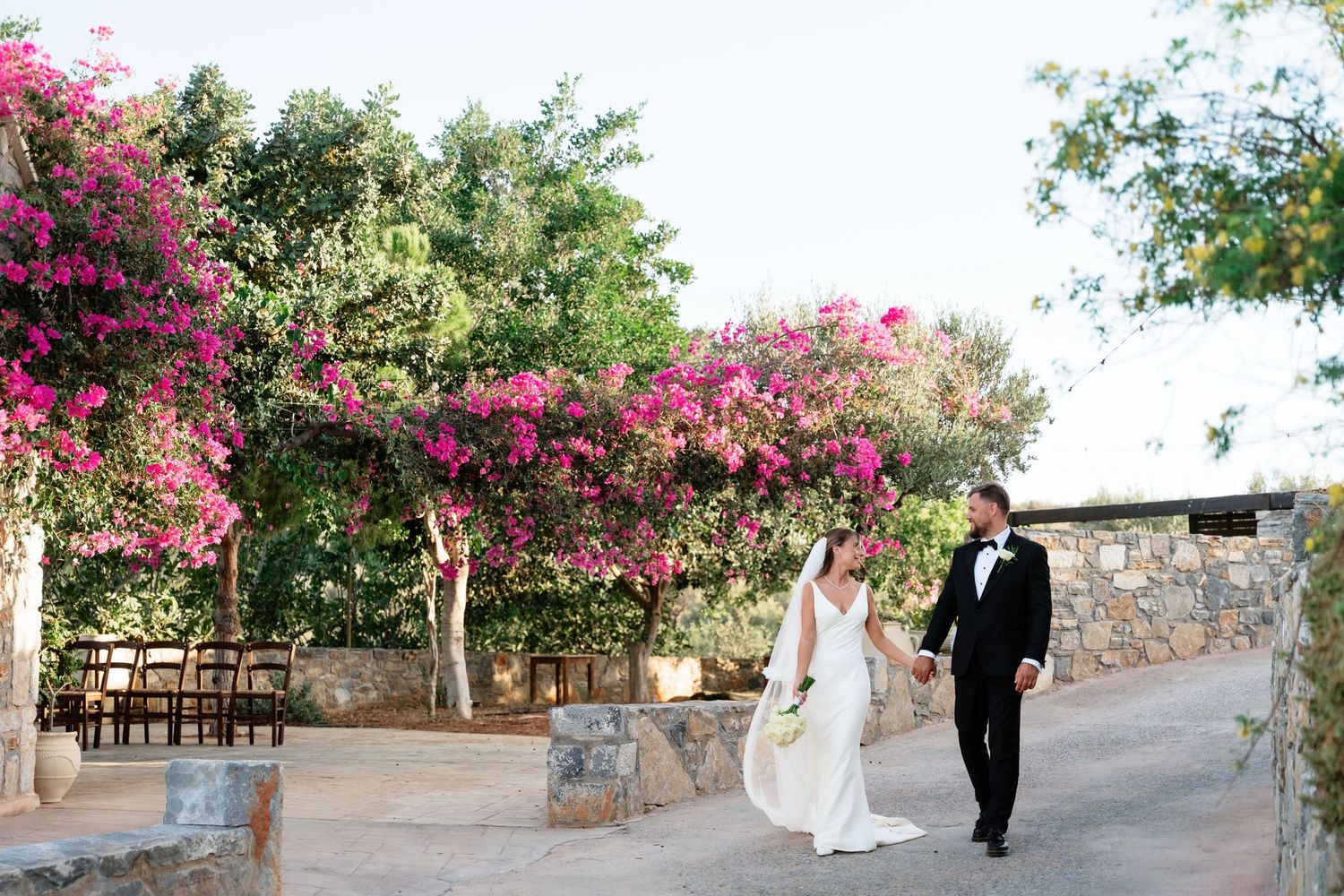 A couple walks together under blooming pink bougainvillea flowers along a stone pathway during a wedding ceremony.