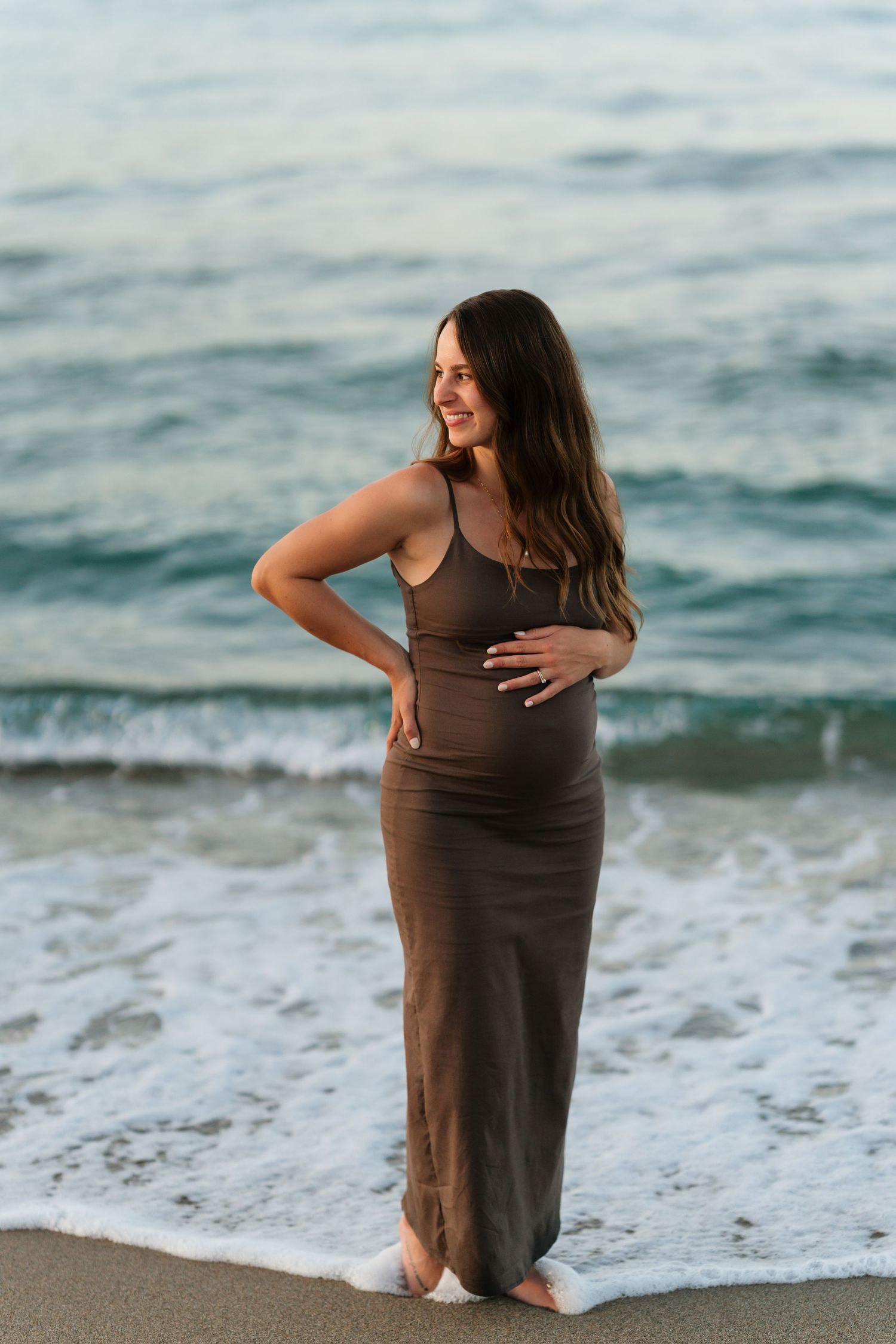 Person in brown dress sits at ocean's edge as waves roll in during sunset at the beach.